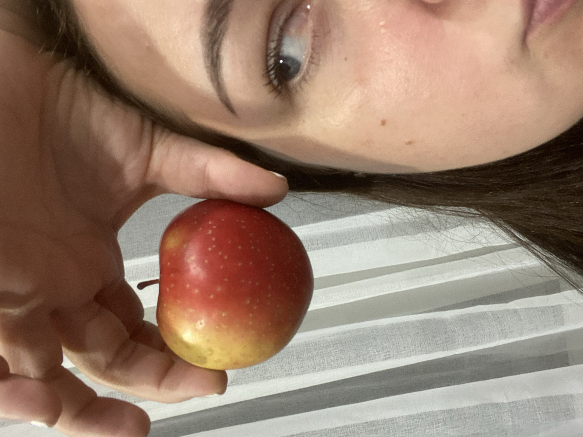 A person with fair skin and brown hair is holding a red and yellow apple in their hand. The background features a white, horizontally striped surface, possibly a window covering or blind. The focus is on the apple and the person's face, with the eye visible and looking upwards. There is no evidence in the image to suggest it was taken in Sochi, Russia.