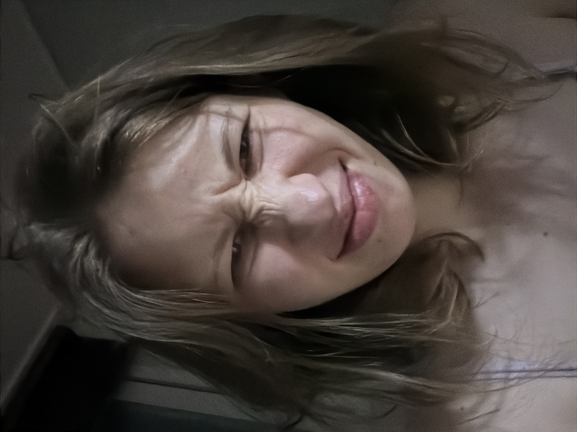 A close-up, low-angle, slightly grainy selfie of a young woman with fair skin, light brown hair, and a wrinkled nose and squinting eyes. She is wearing a light-colored top with thin straps. The background is dark and indistinct, suggesting an indoor setting with low lighting. No specific location in Dongducheon-si, South Korea, can be identified from the image.