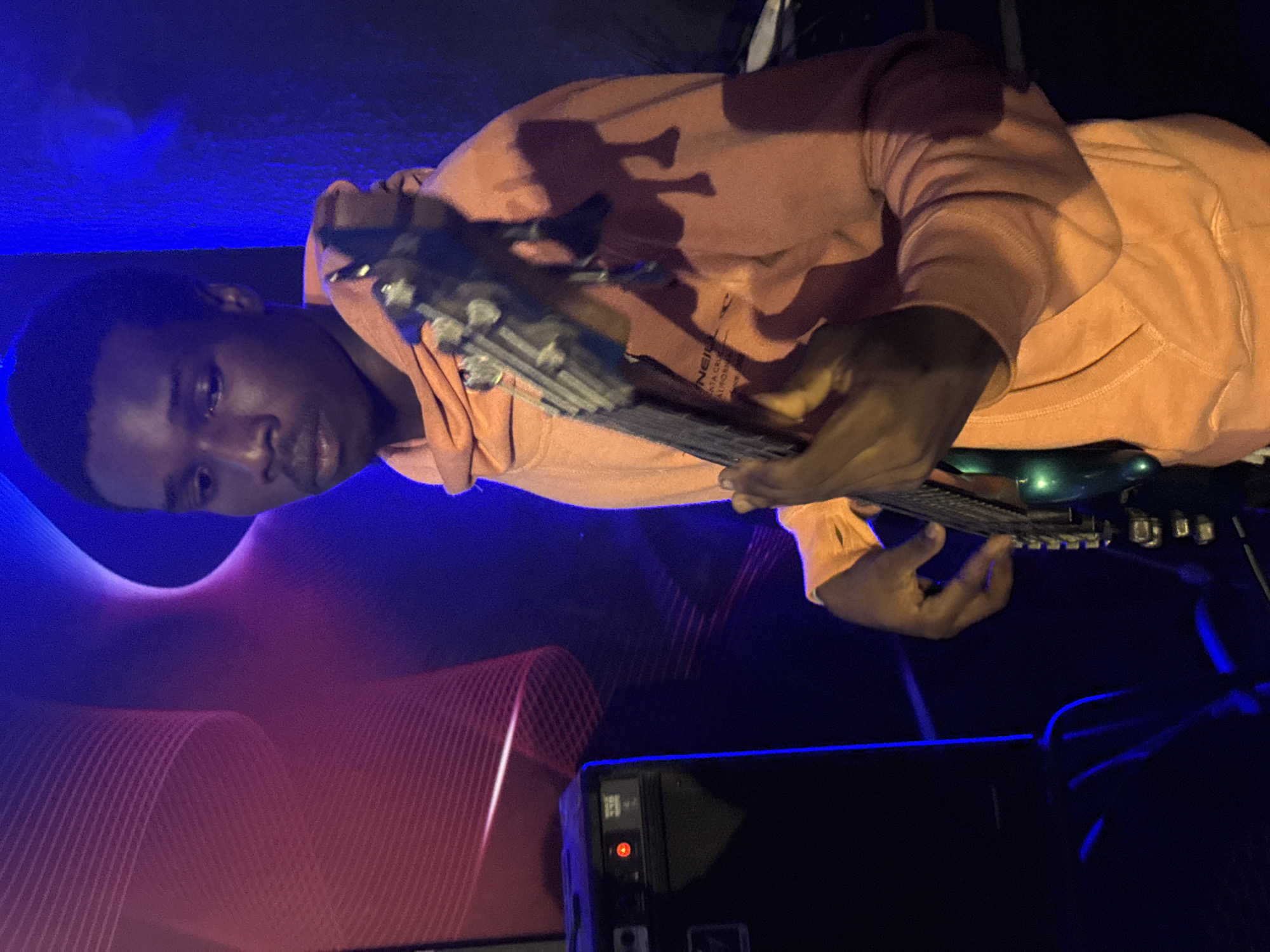 A young Black male is shown from the chest up, playing a bass guitar in a dimly lit room. He is wearing an orange hoodie and appears to be in a performance or practice space. The background features a vibrant blue and red light display, with a graphic wave pattern. To the left of the frame, a black speaker cabinet with a red indicator light is visible. The scene is suggestive of a musical performance or rehearsal, possibly in a venue or studio. There is no information in the image to suggest the location is Nkpor, Nigeria.