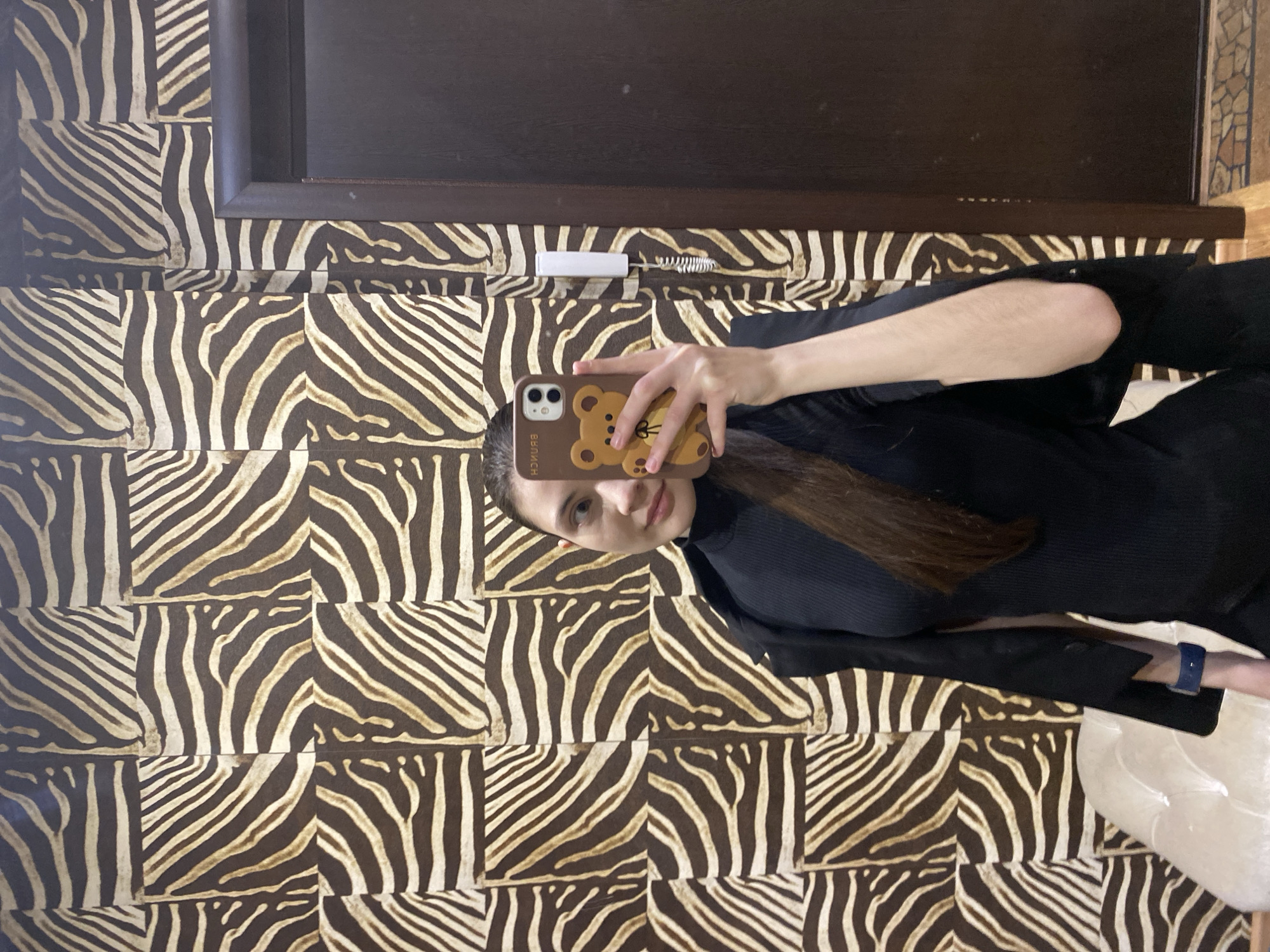 A young woman takes a mirror selfie in an indoor setting, likely in Votkinsk, Russia. She is shown from the chest up, wearing a dark top, and has long, dark brown hair. Her face shows a neutral to slightly smiling expression as she looks towards the phone screen. In her right hand, she holds a smartphone with a light brown case featuring a cartoon bear design and the word "BRUNCH" visible in reverse text. A blue-banded smartwatch is worn on her left wrist.
The background of the mirror reflection is dominated by a distinctive wall covering composed of square panels with a brown and beige zebra stripe pattern. Above this patterned wall, a dark brown wooden frame, possibly part of a door or furniture, spans the upper section of the reflection. A small white electronic device with a coiled wire is mounted on the wall between the wooden frame and the zebra pattern. A blurry, light-colored object is partially visible in the bottom right corner, possibly another piece of furniture or decor. The lighting is consistent with an indoor environment.
