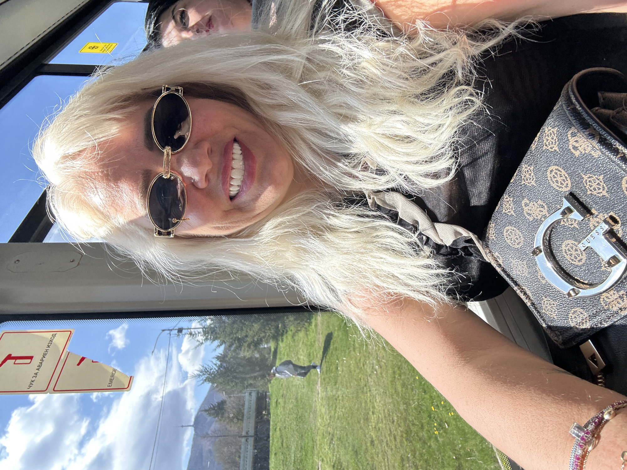 A blond woman wearing sunglasses is in a bus, likely a public bus in Sofia, Bulgaria. The reflection in the window shows a green lawn and a mountain in the distance. Inside the bus, there's an "Emergency Exit" sticker. She is carrying a Guess brand handbag. Another person is partially visible in the background.