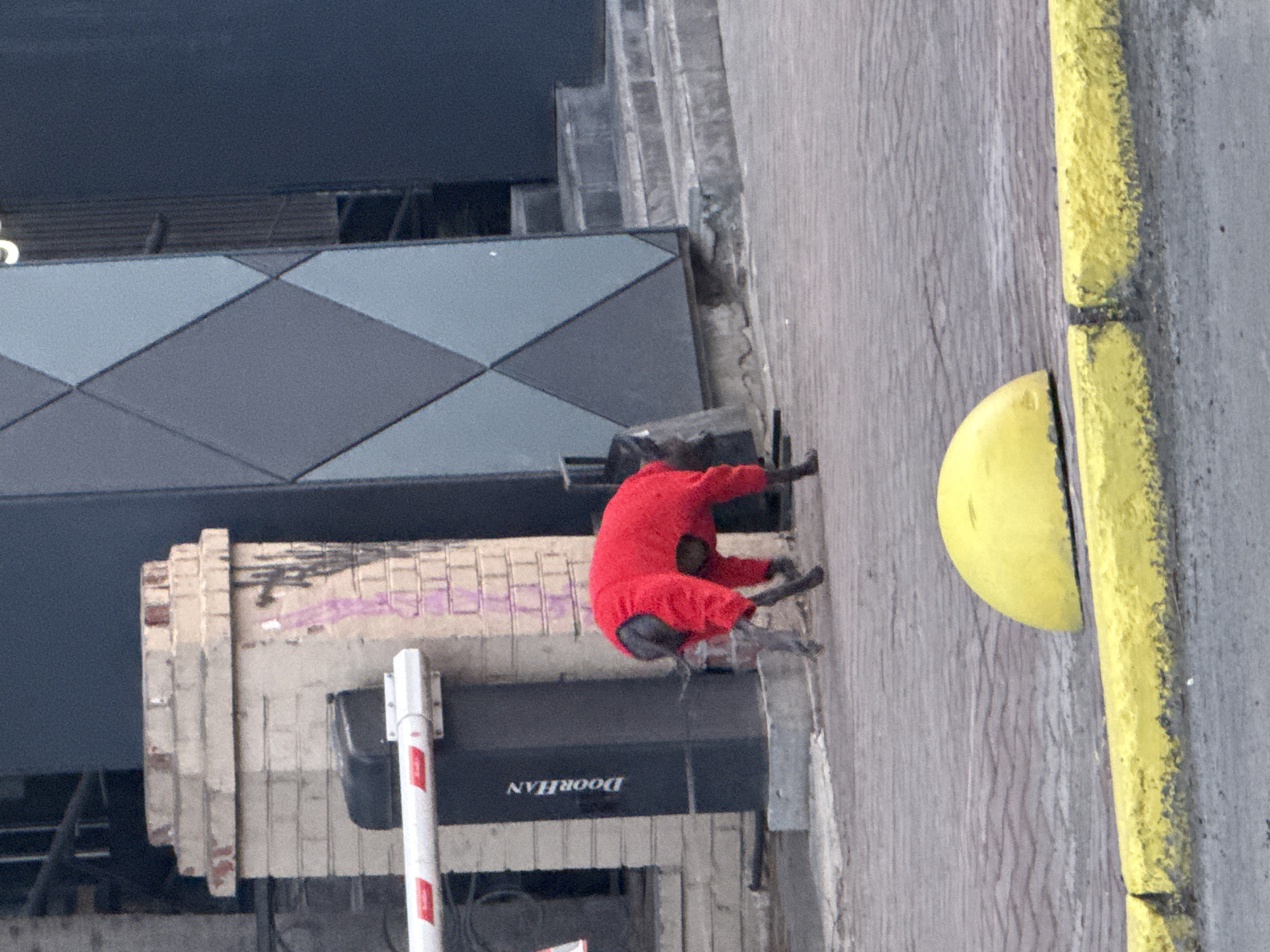 The image shows a person wearing a red jumpsuit and what appear to be black rubber boots climbing a wall in an urban environment. The wall is made of light-colored bricks and is adjacent to a "DOORHAN" branded gate operator. To the right of the person is a yellow-painted curb. Above the person is a building with a geometric pattern of grey diamonds. The person's actions and intent are unclear, but they appear to be in the process of scaling the wall. The location is designated as Unknown, Unknown.