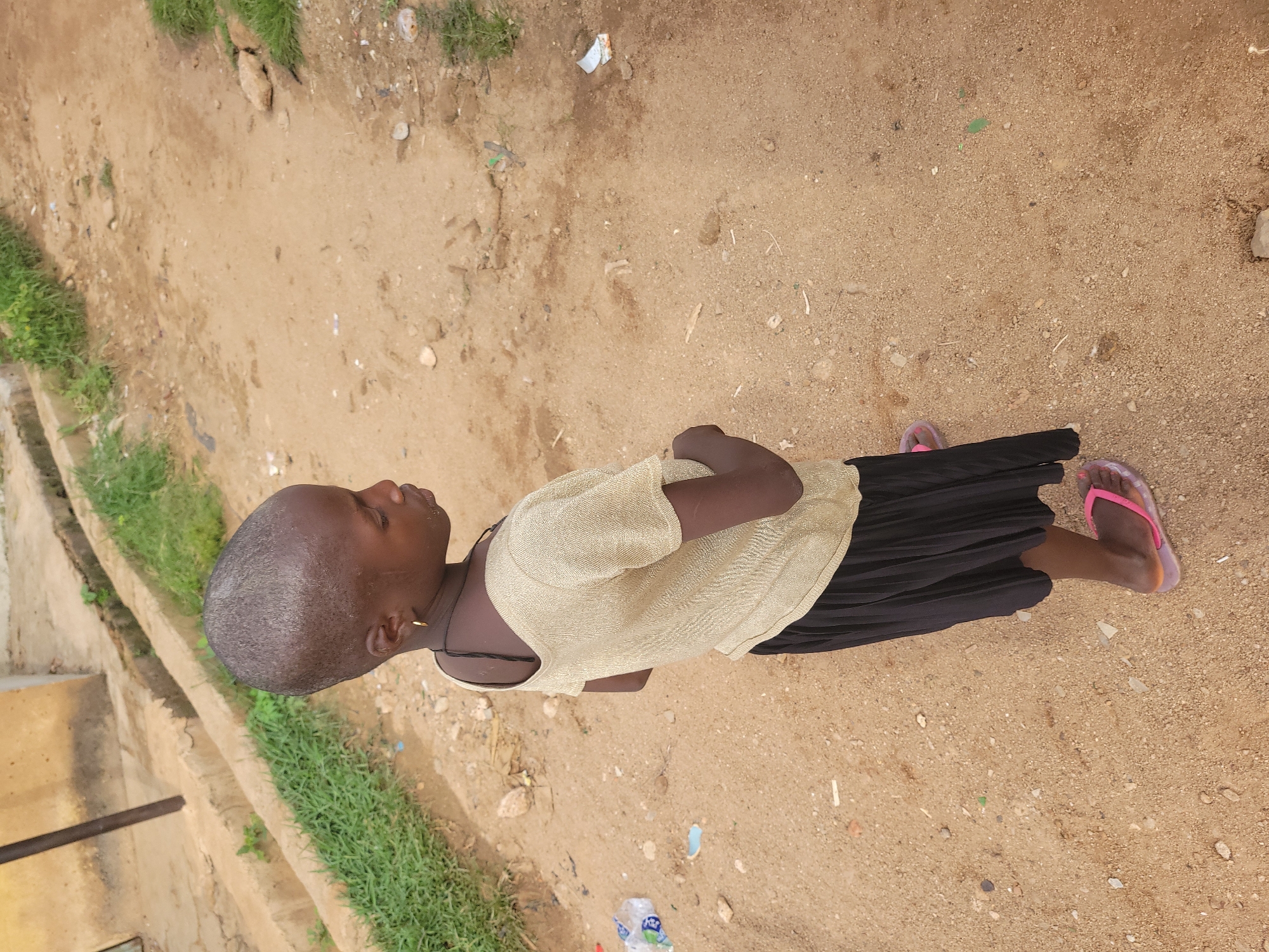 A young girl with a shaved head, wearing a gold-colored sparkly t-shirt and a black pleated skirt, stands on a dirt path. She has a single gold hoop earring in her left ear and a dark string necklace. She is wearing pink flip-flops. The path is uneven and dusty, with scattered small stones and some green vegetation visible on the sides. In the background, there's a raised concrete structure with some water drainage and a glimpse of a building. The overall environment appears to be an outdoor area, possibly a residential or unpaved street within a city.