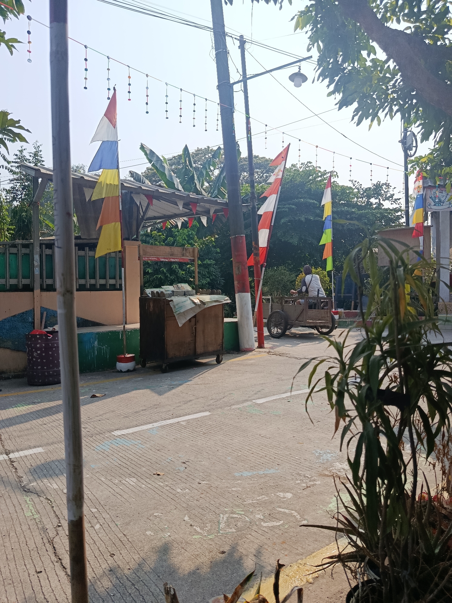 A street scene in Wanasari, Indonesia, captures a bright, sunny day with several elements suggesting a festive or celebratory atmosphere. The foreground is dominated by a concrete road marked with white lines and some scuff marks. A tall, grey-and-white striped pole is positioned to the left, with a dark cylindrical object with a red lid at its base. Further back on the left is a low wall with green and pink sections.
A wooden cart on wheels, used for vending, is placed in the middle of the scene, covered with a light-colored tarp. Behind this cart, a person is seated on a similar wooden cart with two wheels, facing away from the camera and appearing to be moving. This person is wearing a white shirt.
Decorations are prominent throughout the image. Several Indonesian flags, red and white, are displayed on poles. Additionally, colorful triangular pennants are strung overhead, and there are strings of small, colorful circular ornaments hanging between poles. Trees with dense green foliage are visible in the background, and the sky is clear and bright. The overall impression is one of an outdoor market or a public space decorated for an event.