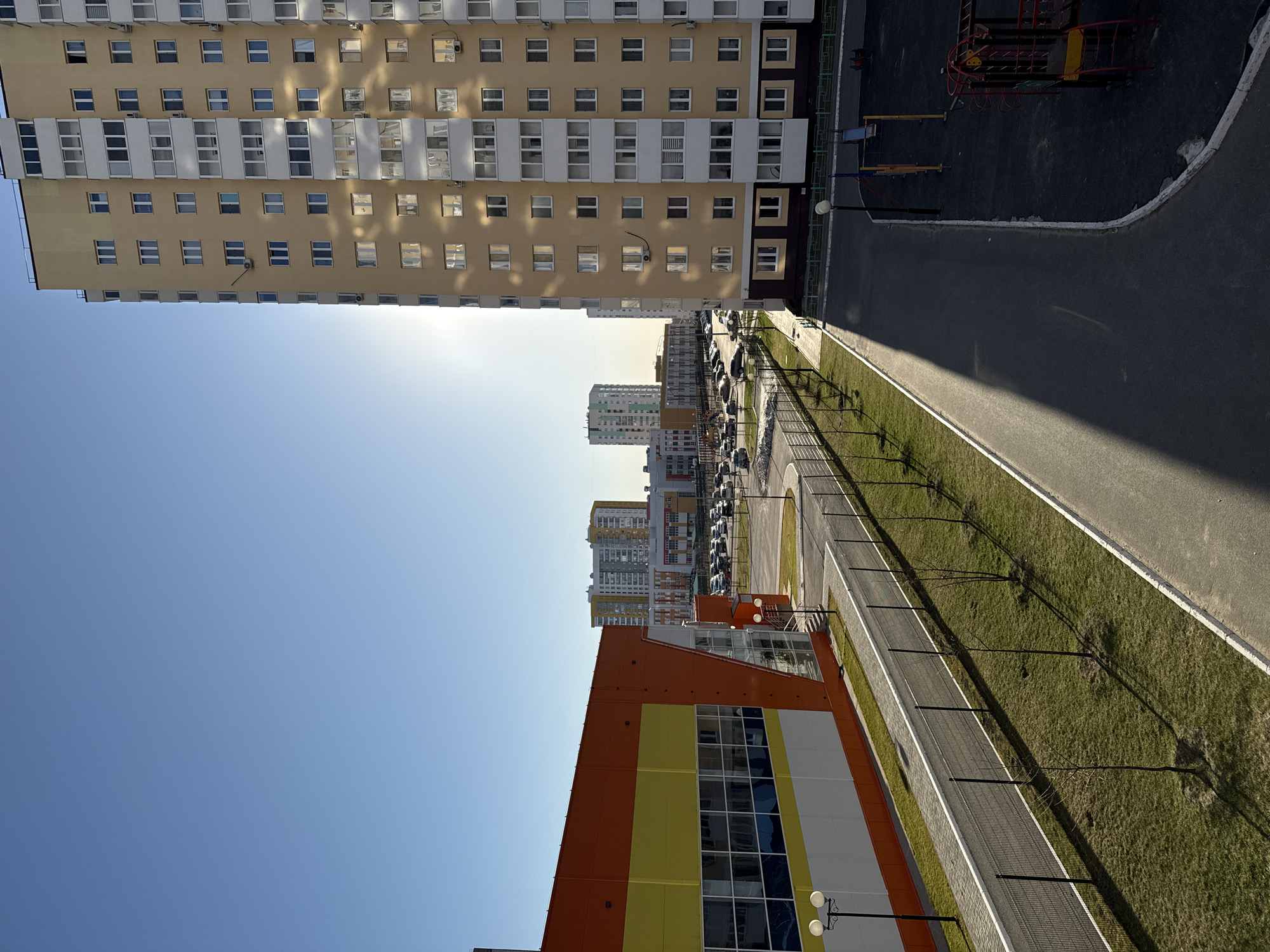 The image depicts a cityscape in Tobolsk, Russia, featuring modern residential and commercial buildings under a clear blue sky. A tall apartment building with rows of windows dominates the upper left quadrant. Adjacent to it is an asphalt area with a playground. In the distance, other multi-story buildings are visible. A lower-level building with orange, yellow, and white panels stands in the lower portion of the frame, alongside a paved walkway lined with young trees and lampposts. A parking lot is visible beyond the walkway. There are no people visible in the image.