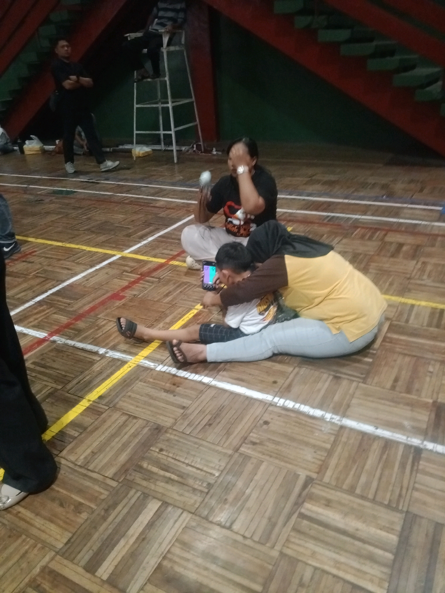 The image depicts an indoor wooden floor court with line markings, possibly a sports facility. In the foreground, a person in a black outfit with a white stripe on their pants and grey sneakers stands with arms crossed. Behind them, a white step ladder structure is visible with a person seated on it. Further back, a woman in a black t-shirt is seated on the floor, covering her eyes with her hand. To the right, a person wearing a yellow shirt and grey pants is seated on the floor, embracing another person who is leaning on them and looking at a smartphone. The scene appears to be within a building in Wuryorejo, Indonesia.