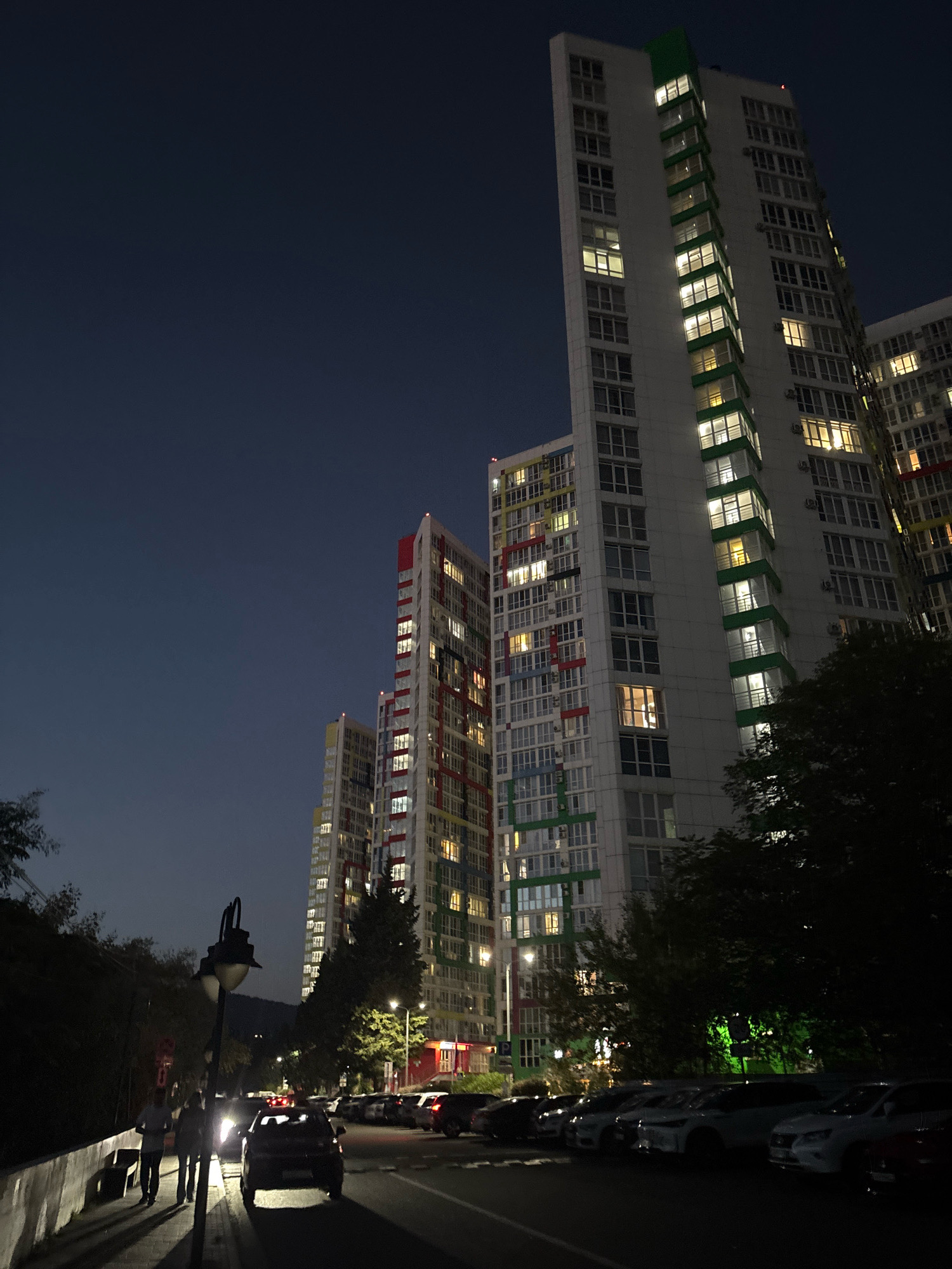 The image depicts a nighttime street scene in Sochi, Russia, characterized by modern high-rise residential buildings with illuminated windows. In the foreground, a street lamp casts shadows across a sidewalk where two individuals are walking away from the viewer. A dark-colored car is parked on the right side of the street, with its headlights on. Beyond this, several other cars are parked in a row. The sky is a dark blue, indicating it is evening or night. Trees line the left side of the street, creating a stark contrast with the brightly lit buildings. The architecture of the apartment complexes suggests a relatively new urban development.