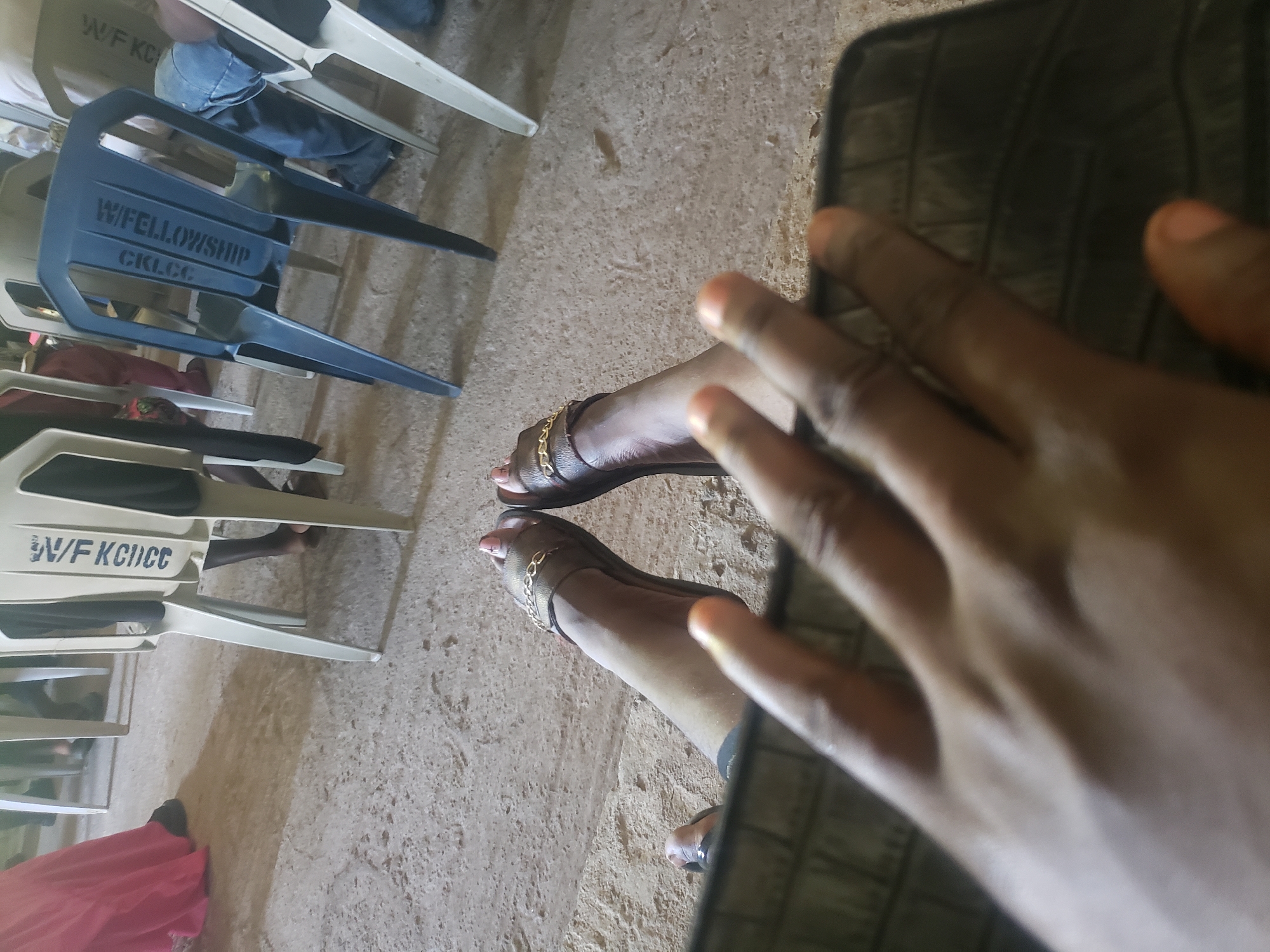 The image depicts an indoor scene in Narayi, Nigeria, captured from a low, first-person perspective. In the immediate foreground, a blurred, dark-skinned right hand partially obscures a dark, textured object, likely a mobile device or a book/binder cover. The photographer's dark-skinned lower legs and feet are extended forward, wearing brown flat sandals with gold-colored chain embellishments. The toenails are manicured with light pink polish, appearing in a French tip style.
In the mid-to-background, multiple individuals are partially visible, seated on plastic chairs. A person on the left is seen wearing a long pink garment and dark footwear. Another individual on the right is dressed in blue jeans. Several plastic chairs are arranged in rows. One off-white chair displays "W/F KGICC" printed vertically in dark blue text on its backrest. Another dark blue chair prominently features "W/FELLOWSHIP" and "GKLC.CO" printed vertically in light text. A third off-white chair is partially visible on the right, also bearing "W/F KCICC". The flooring is a light brown, textured surface consistent with concrete or unpolished tile, showing signs of wear and minor scattered debris. The overall environment suggests a communal gathering or meeting.