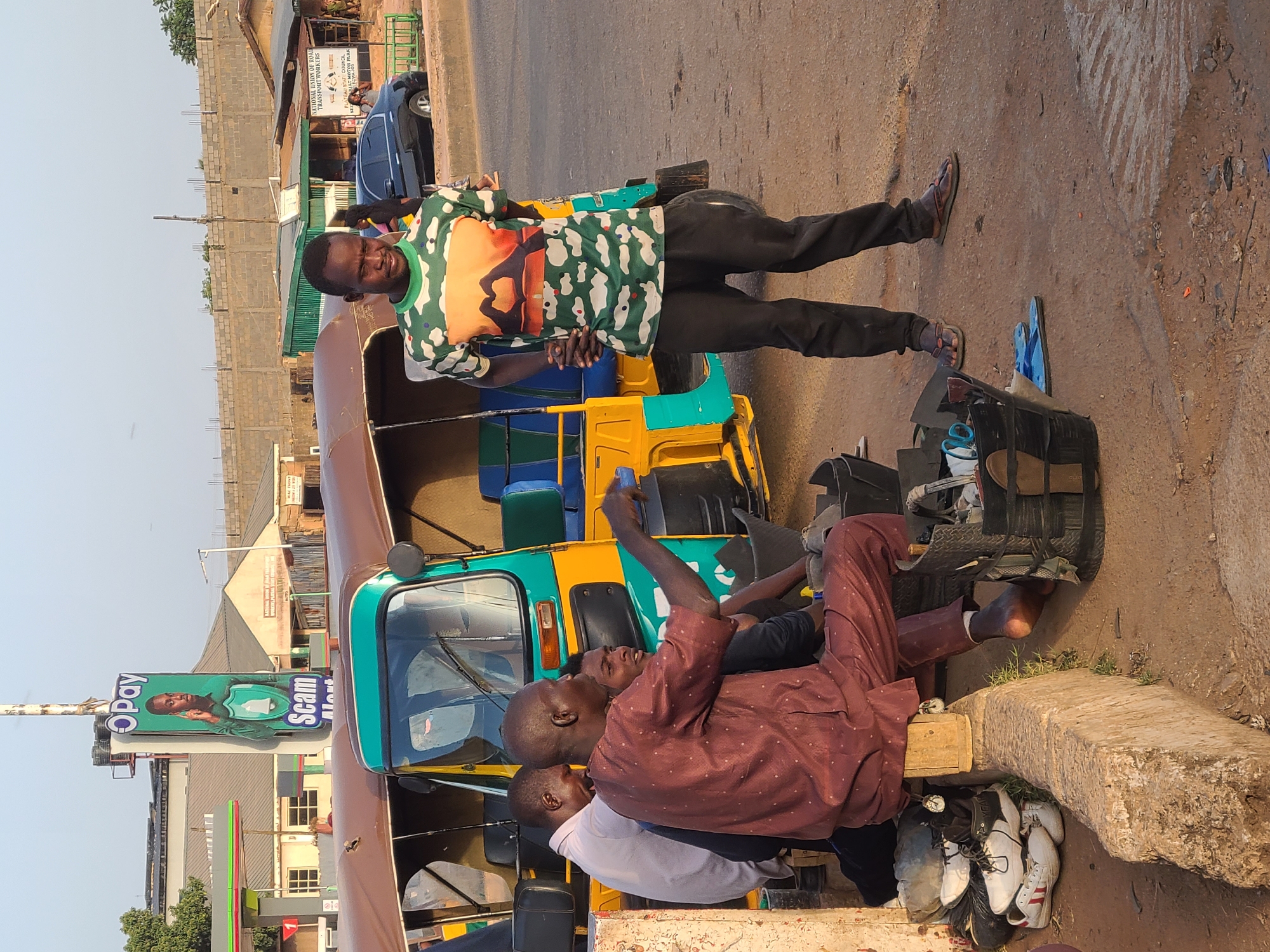 The image captures a vibrant street scene in Jos, Nigeria, depicting a group of men engaged in daily activities under clear, bright daylight. The scene is centered around a three-wheeled vehicle, commonly known as a keke napep or auto-rickshaw, which is teal, yellow, and brown.
In the foreground, a man in a maroon long-sleeved shirt and trousers sits on a concrete block, holding a mobile phone, while another man (partially visible, wearing a white shirt) appears to be attending to his shoes, suggesting a shoeshine service. A young boy is also visible between them. Next to this interaction is a shoeshine kit crafted from wood and a tire, surrounded by several pairs of shoes and sandals.
Standing prominently to the right of the keke napep, a man smiles directly at the camera. He wears a distinctive green, orange, and white patterned short-sleeved shirt over black trousers and flip-flops.
The background reveals a bustling urban environment with various buildings, including brick structures, some under construction, and others with corrugated roofs. A road or dirt path runs through the scene. On the left side of the image, a vertical billboard prominently displays "Opay Scam Alert" with an image of a man and a shield icon. Further in the background, a less distinct sign reads "NATIONAL UNION OF ROAD TRANSPORT WORKERS" and "JOS LAL ESTATE COUNCIL MOTOR PARK," indicating a transport hub.
The overall atmosphere is lively, characteristic of a typical day in a Nigerian city or town, with people going about their business.