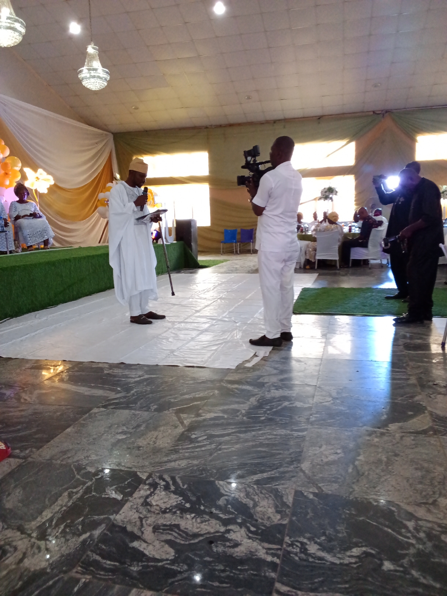 A man in a white Islamic robe and cap stands on a white sheet laid over a tiled floor, holding a microphone and reading from a paper. He is facing left, away from the camera, and has a walking stick planted beside him. Behind him, a man in a white shirt and trousers operates a video camera. Further back, several people sit at tables, with two men in black standing closer to the camera. The environment appears to be an indoor event space decorated with white and gold drapes and illuminated by chandeliers. The floor is a dark marble.