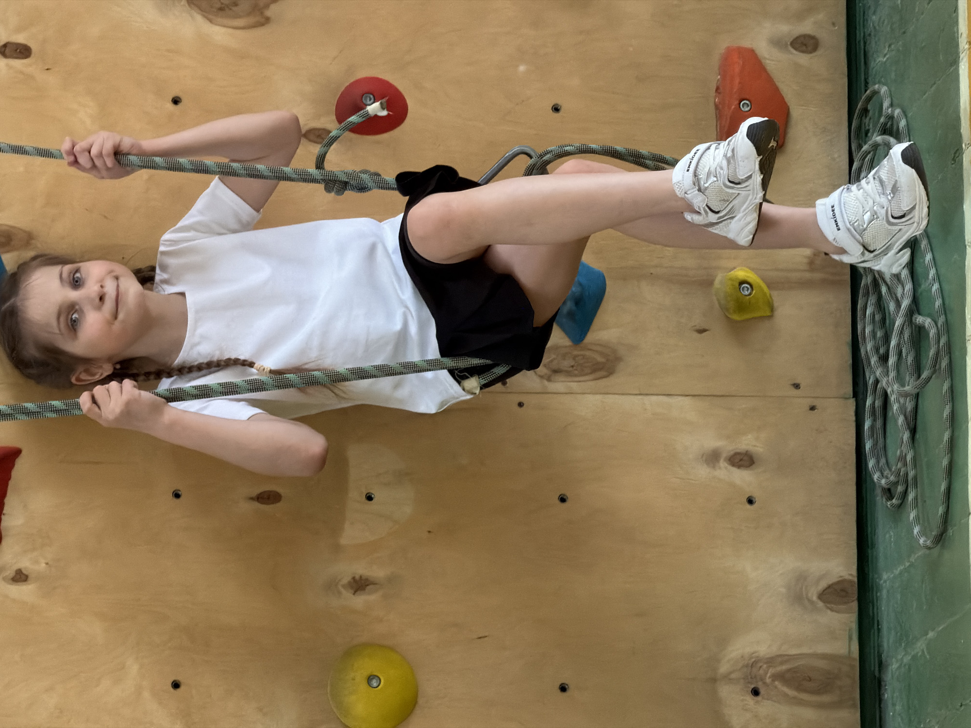 A young girl with light brown braided hair and blue eyes is captured in a horizontal orientation on a wooden climbing wall. She is wearing a white short-sleeved t-shirt, black shorts, and white sneakers. Her body is suspended parallel to the wall, held by a green and white patterned climbing rope which she grips with both hands. A carabiner or part of a harness is visible near her waist, connecting her to the rope.
The climbing wall is made of light-colored plywood with various colored climbing holds attached, including red, orange, yellow, and blue. Her feet, clad in white athletic shoes, are bent at the knees, and the brand name "ENKJOYE" is visible on the side of her right sneaker. One orange hold is visible near her right foot, and a yellow circular hold is beneath it.
The girl has a slight smile and wide eyes, indicating focus, enjoyment, or playfulness during her activity. The setting appears to be an indoor climbing gym or a recreational area, possibly located in Izhevsk, Russia. The overall impression is one of active engagement in a climbing or training exercise.