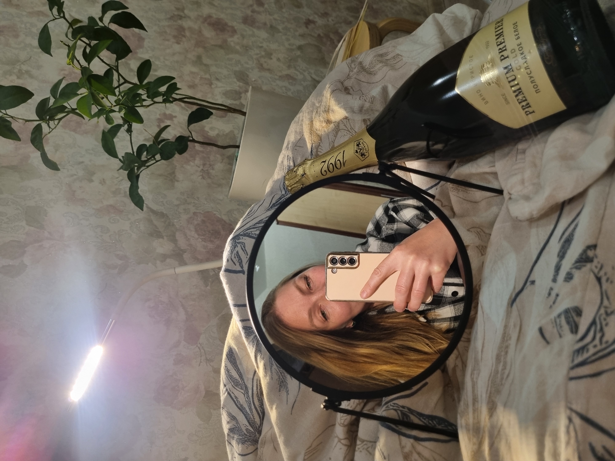 A person is taking a selfie in a round mirror. The person has long brown hair and is wearing a plaid shirt. They are holding a tan-colored smartphone. A bottle of Premium Premier Gold wine is lying on a patterned beige blanket. A plant is visible in the upper left of the image. A desk lamp shines brightly in the lower left of the image. The location is likely Chaikovsky, Russia based on the Cyrillic writing on the wine bottle label.