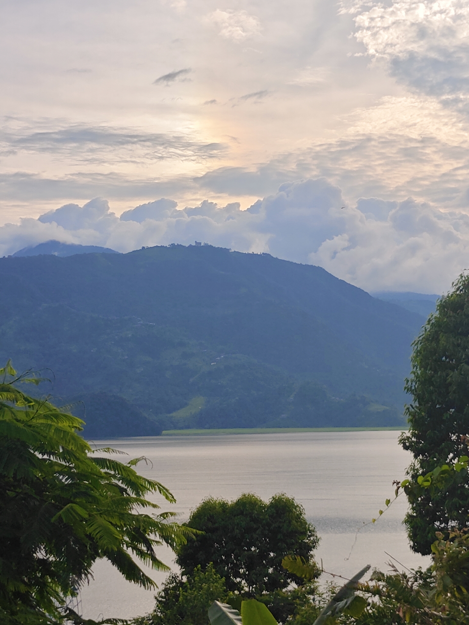 This is a landscape photograph taken in Pokhara, Nepal, overlooking the Phewa Lake. The foreground features lush green foliage of trees and plants, with some of their leaves and branches obscuring parts of the view. In the midground, the calm waters of Phewa Lake stretch across the frame, reflecting the soft light of the sky. Beyond the lake, a series of rolling hills and mountains rise, covered in dense greenery. The higher peaks are partially shrouded in clouds, creating a layered effect. The sky above is a blend of soft whites, grays, and hints of pale yellow, suggesting either early morning or late afternoon. The overall impression is one of serene natural beauty.