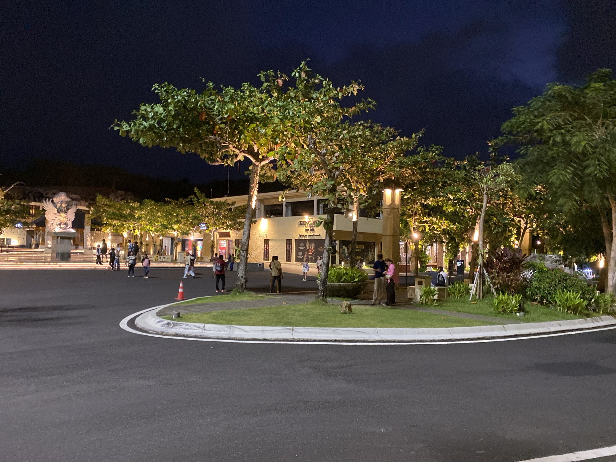 The image depicts a nighttime scene in Kuta Selatan, Indonesia, likely a public square or promenade. In the foreground, a paved road curves from the bottom left to the right, with a white-lined curb and a grassy median. The median features trees with illuminated foliage, some with red fruits, and streetlights. Several people are visible standing and walking on the paved area behind the median, some near buildings and others in a more open space.
To the left of the scene, a large stone sculpture or monument with outstretched wings is visible, illuminated by lights. Behind the sculpture and extending across the mid-ground are low-rise buildings with illuminated windows and signs, suggesting shops or restaurants. The architecture appears to be open-air or with large storefronts.
Several trees with broad leaves are interspersed throughout the scene, some illuminated from below, casting warm light on their branches and foliage. There are also bushes and other greenery in the median and around the buildings. A traffic cone with an orange top is placed on the grassy median near the curb. The sky is dark, indicating nighttime, with some clouds visible in the upper right portion of the image. The overall atmosphere is that of a well-lit public gathering space at night.