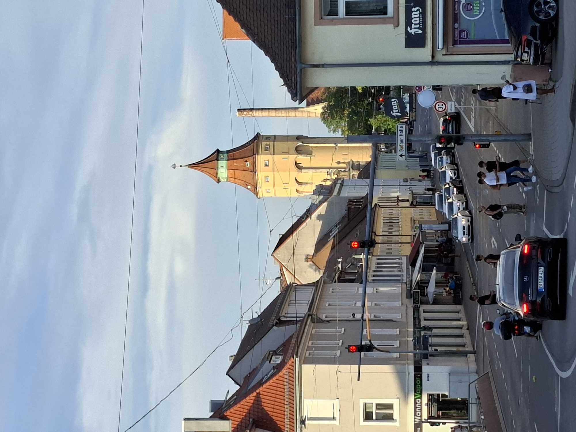 The image captures a street scene in Rastatt, Germany, featuring a prominent water tower in the background. The perspective is from street level, looking down a road lined with buildings. The sky is partly cloudy, with visible overhead wires crossing the scene.
In the foreground, a black sedan with license plate "RA AA 417" is stopped at a traffic light. A person is riding a scooter behind the sedan, also stopped. Several pedestrians are visible crossing the street near the intersection. To the right, a woman in a white dress stands on the sidewalk next to a black car.
Along the street, various businesses are present, including "Wanna Vapor" on the left and "Franz Bastatt" on the right, with a "Kiosk" sign visible within the latter's window. Several other cars are seen on the road, either stopped at the traffic light or in motion further down the street. People are seen walking on the sidewalks and seated at outdoor tables of a cafe.
The iconic yellow water tower with a red conical roof dominates the middle ground, with a tall, slender smokestack visible behind it. The buildings lining the street are a mix of residential and commercial, with red-tiled roofs visible on some. Traffic signals are active, showing red lights at the intersection. A street sign indicates "Badner Halle." A speed limit sign displaying "30" is also visible. The overall atmosphere suggests a typical late afternoon or early evening in a European town.