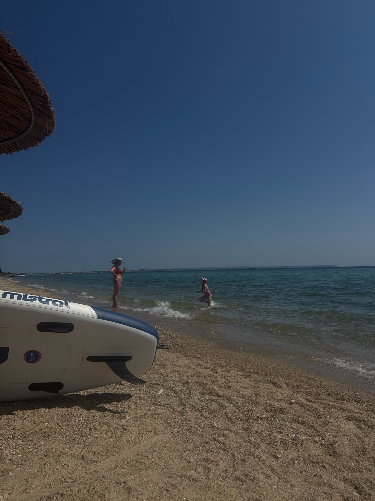 The image depicts a sunny day at a beach in Nea Moudania, Greece. In the foreground, a white and blue inflatable paddleboard rests on the sand. To the left, a straw umbrella provides shade. In the middle ground, a woman in a red bikini and a straw hat stands in the shallow water, holding a mobile phone. Further to the right, a child in a pink swimsuit runs towards the sea. The sea is calm and blue, with a clear horizon. The beach itself is sandy, with some small pebbles. The sky is a vibrant, cloudless blue.