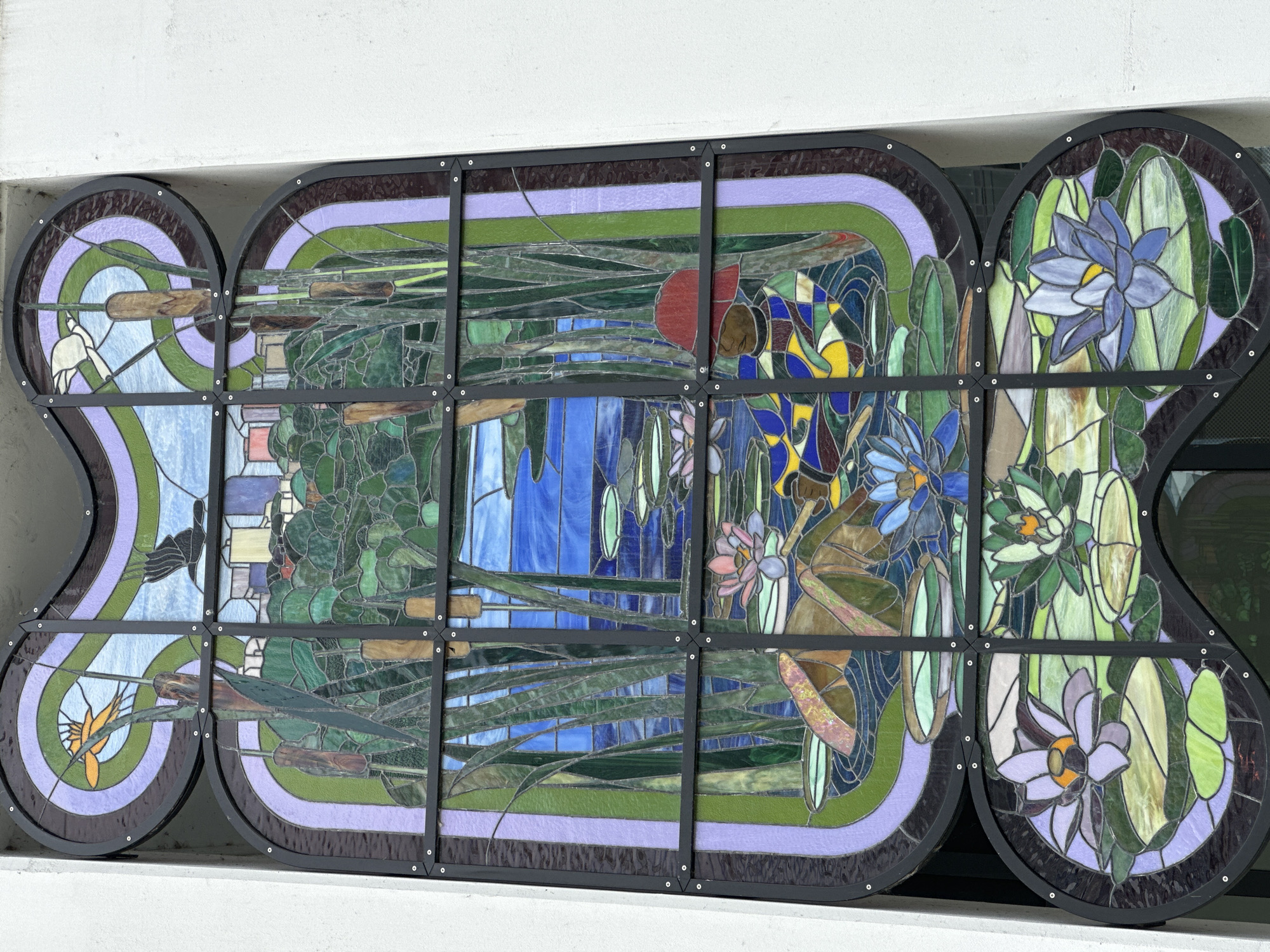 The image shows a stained glass window depicting a serene pond scene with water lilies, reeds, and a distant cityscape. The window is installed on the exterior wall of a building. In the upper portion, two birds are depicted in flight against a sky of swirling blues and purples. Below this, a lush green landscape with a body of water stretches across the window. Reeds and cattails frame the central scene, with lily pads and water lilies floating on the water's surface. A figure wearing a red hat appears to be interacting with the pond. The lower section of the window features more water lilies in bloom. The overall impression is one of nature and tranquility. This stained glass window is located at the Burnaby Public Library - McGill Branch in Burnaby, Canada.