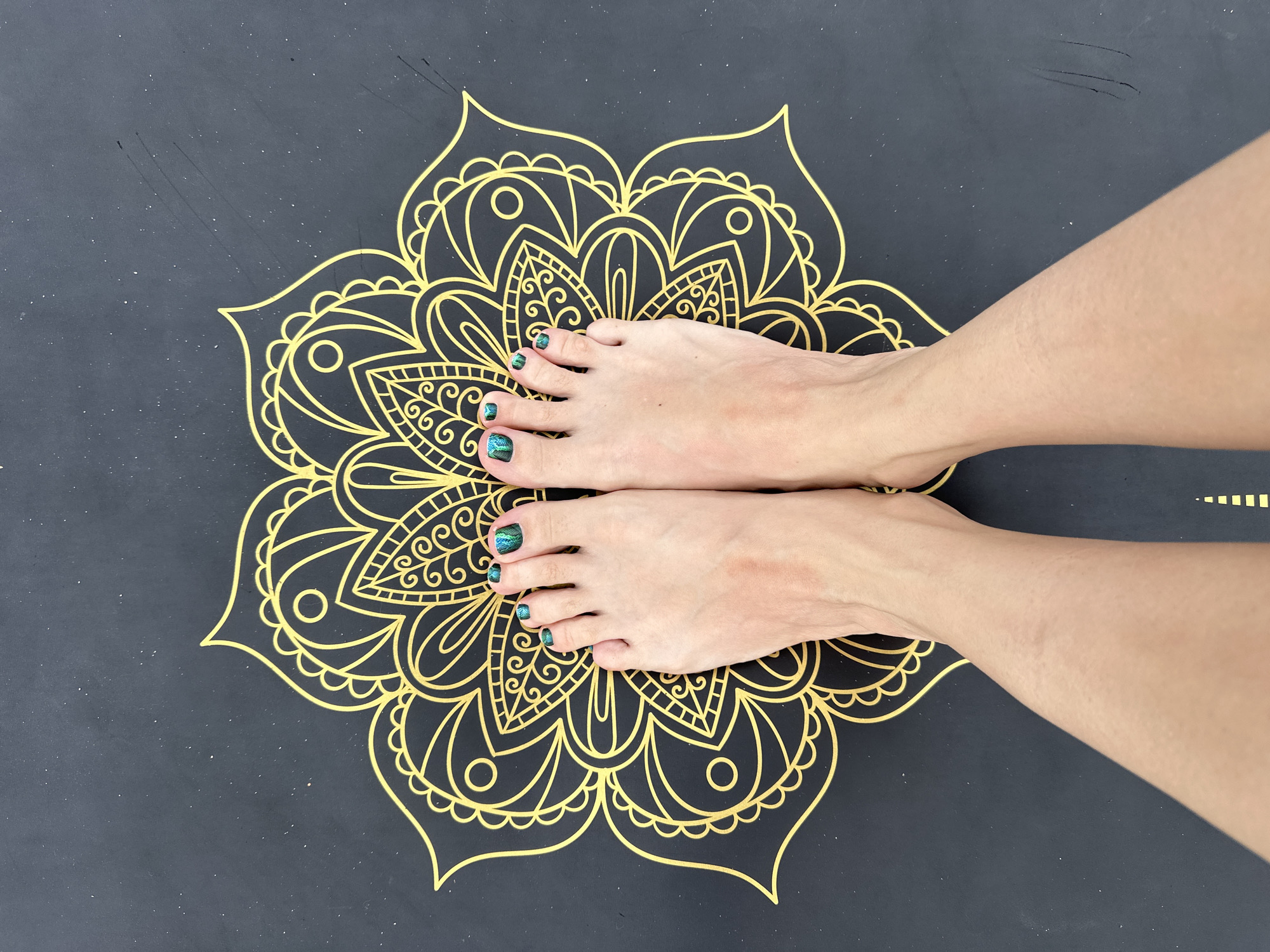 A pair of bare feet with fair skin is prominently displayed in a top-down view, resting on a dark-colored surface, likely a yoga or exercise mat. The toenails are painted with a striking, iridescent teal-green metallic polish. Beneath the feet, a large, intricate mandala design in metallic gold is centered on the dark mat, featuring symmetrical floral and geometric patterns that radiate outwards. The mat itself appears to be a matte black or dark grey, with subtle speckles and a few light scuff marks visible on its surface. The image captures a moment of stillness, potentially before or after a yoga or meditation session, or simply highlighting the mat's design and the pedicured feet. No activity is actively taking place. There are no discernible location cues within the image, though the context indicates Vladimir, Russia. The time of day and weather are not evident. No visible text is present.