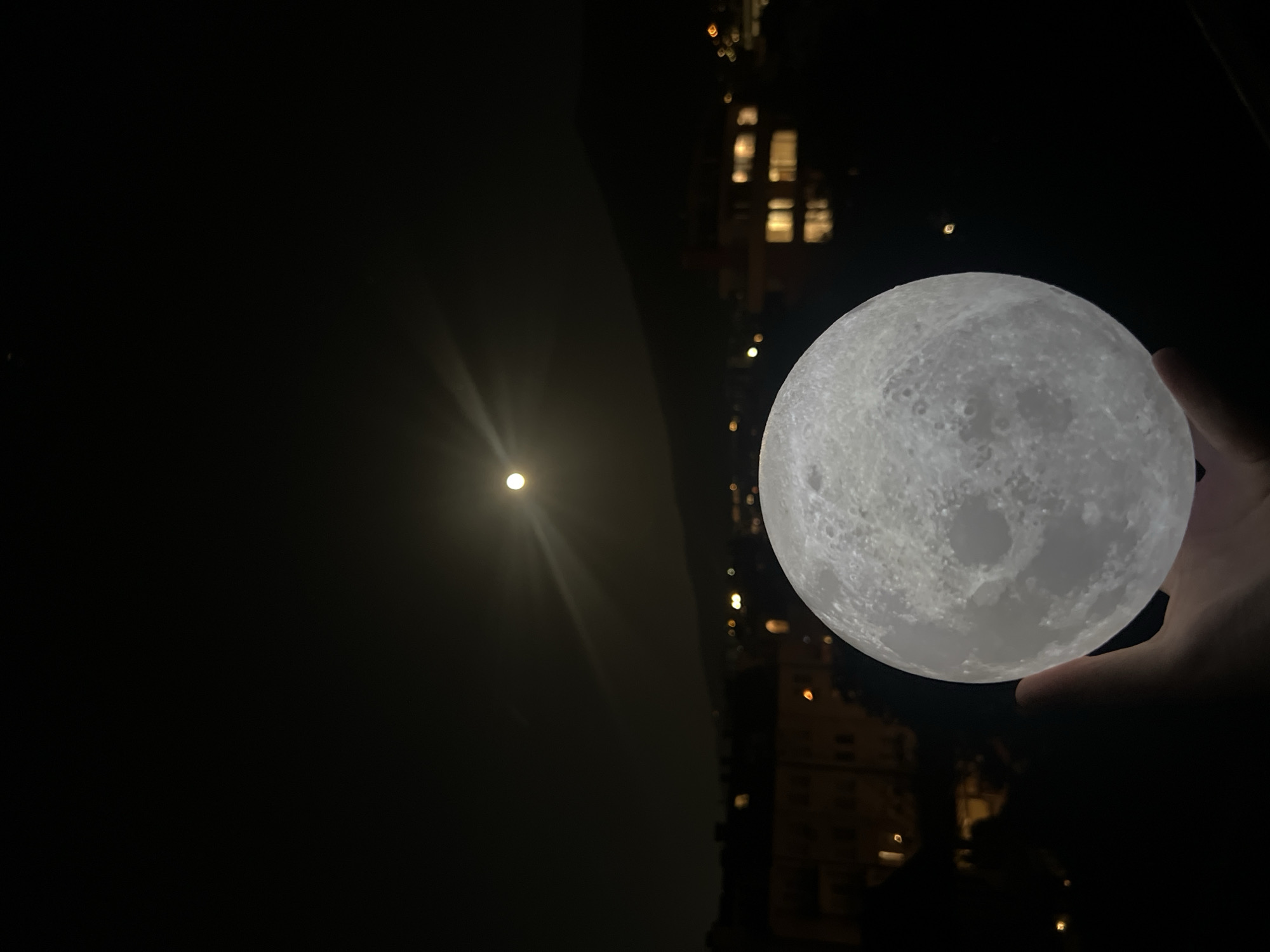 A person's hand holds a spherical, glowing moon-shaped lamp in the darkness. In the background, out of focus, is an actual moon in the dark sky, shining with a distinct glare, and the lights of a city at night, including lit windows in buildings. The location is Sofia, Bulgaria.