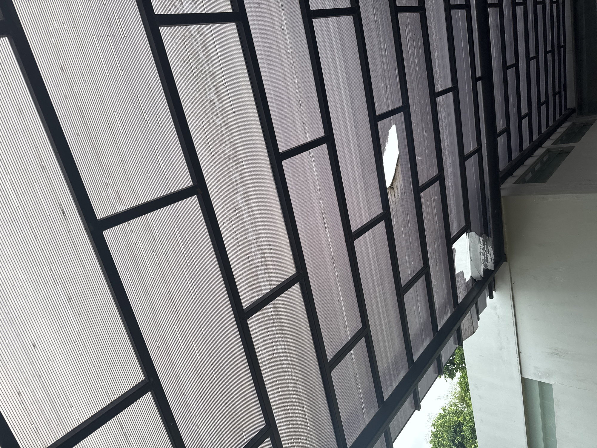 An upward-looking view captures a translucent canopy attached to a light-colored building, likely in Kuta Selatan, Indonesia. The canopy features a dark, possibly black, metal frame forming a grid pattern. Within this grid, ribbed, semi-transparent panels, appearing to be polycarbonate or similar material, allow diffused light to pass through.
Two distinct sections of the canopy show damage, with significant portions of the translucent panels broken or missing. One large, irregularly shaped hole is visible towards the middle-right of the image, while another damaged area appears further to the right, near where the canopy meets the building. These broken sections reveal direct bright light from above.
The panels themselves show signs of weathering or dirt, with some streaking and discoloration. The supporting frame extends into the distance, suggesting a long covered walkway or patio. To the lower right, outside the canopy and adjacent to the building wall, green foliage from trees or bushes is visible, indicating an outdoor setting. The bright light filtering through and from the damaged areas suggests it is daytime. No visible text is present in the image.