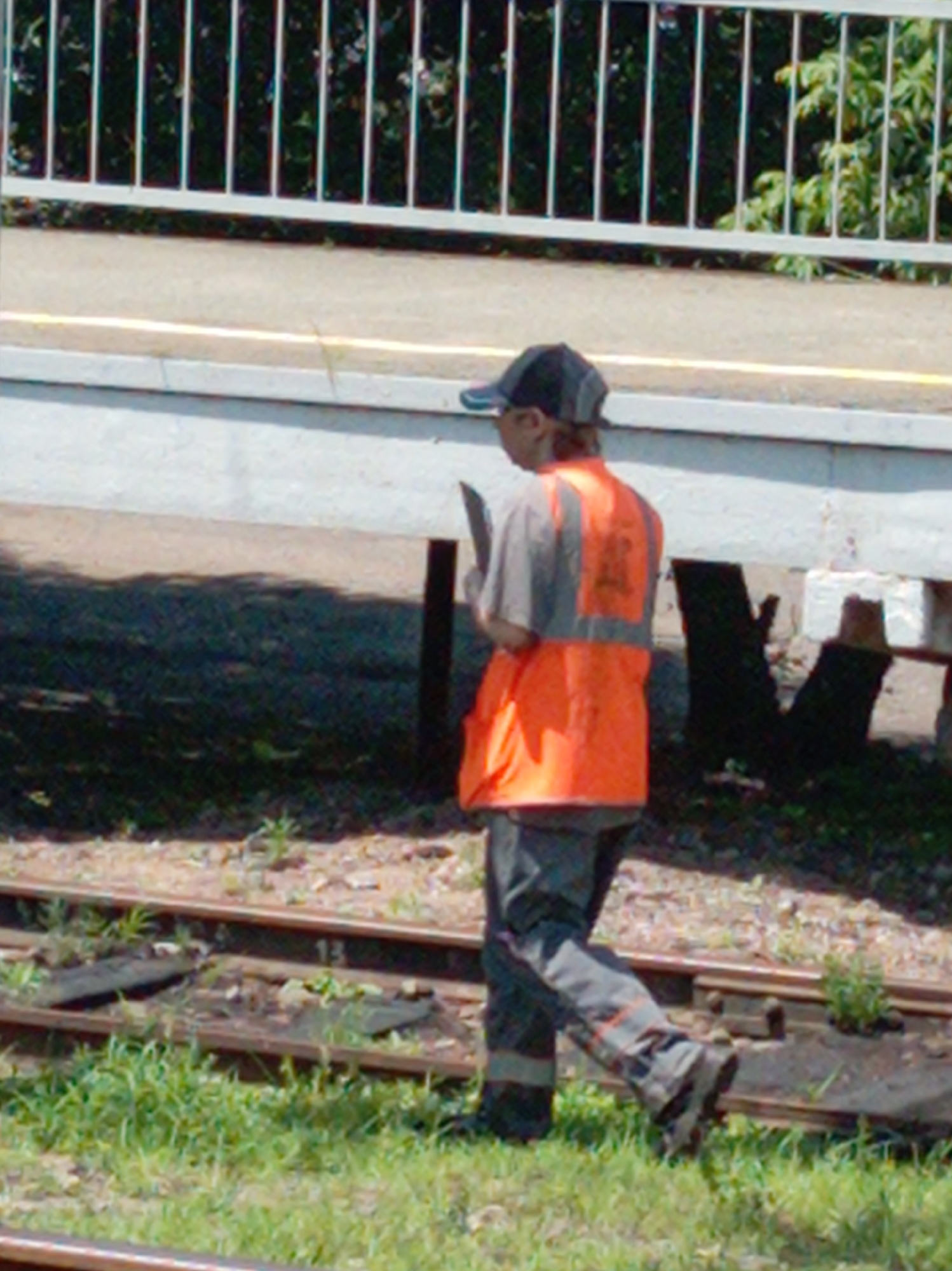 A person wearing a grey cap, a light-colored t-shirt with a grey vest, and grey pants with orange reflective stripes is walking on a dirt and grassy area next to train tracks. The person is also wearing dark boots and a bright orange safety vest over their t-shirt and grey vest. They are holding a dark rectangular object in their right hand, possibly a phone or small device. The background shows a white railing with vertical bars and some foliage behind it. The train tracks are visible in the foreground, with some debris and grass growing around them. The scene appears to be an outdoor industrial or railway area.