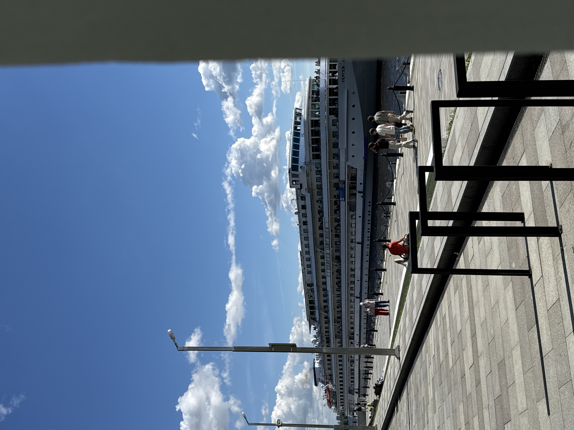 The image captures a sunny day in Khimki, Russia, likely near the Rechnoy Vokzal (River Terminal). A large, white passenger ship with multiple decks is docked along a paved waterfront promenade. The ship's name, "СЕРГЕЙ А..." is visible on its hull. Several people are on the deck of the ship, and a few are visible on the promenade. On the promenade, a man in a red shirt is sitting on a low black structure, facing the ship. Further down the promenade, a couple in casual clothing is walking away from the camera. In the background, a street lamp stands tall. The sky is a vibrant blue with fluffy white clouds, and the sunlight casts shadows on the paved ground. The scene suggests a leisurely atmosphere by the river.