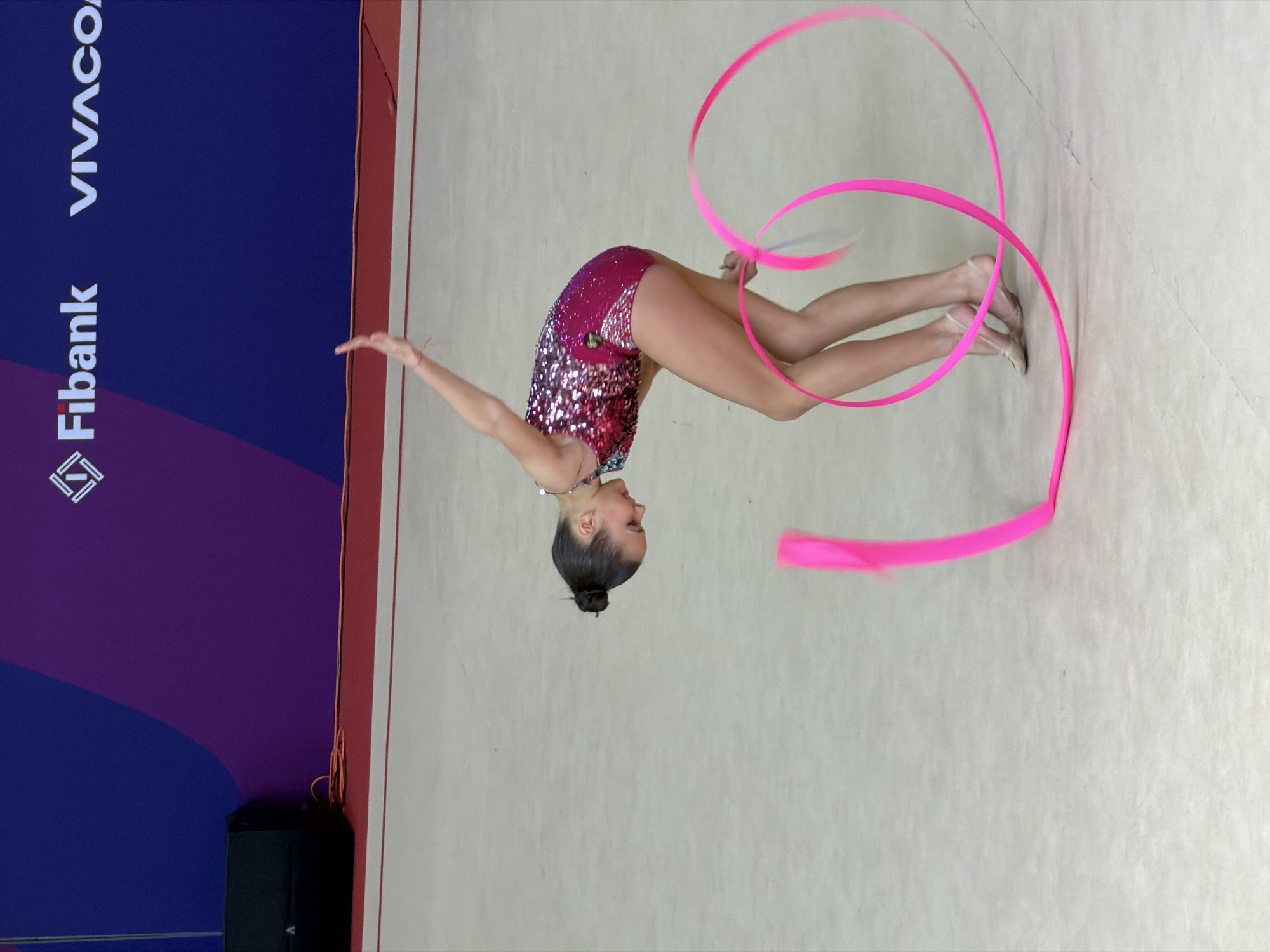 This image depicts a female rhythmic gymnast performing in an indoor arena, likely during a competition or event in Sofia, Bulgaria. The gymnast is captured mid-movement, inverted in a dynamic back arch with her head tilted upwards. She wears a striking, heavily sequined dark pink leotard that shimmers in the light, and her hair is neatly styled in a bun.
A bright pink ribbon is the main apparatus, swirling in elegant loops around her as she performs, with one arm extended towards it and the other reaching outwards. Her legs are extended and pointed, wearing specialized gymnastics toe shoes. The performance takes place on a light-colored, textured mat or floor.
In the background, to the left, is a tall, dark blue panel acting as a backdrop or advertising board. This panel features white sponsor logos: "Fibank" and "VIVACOM" are clearly visible. A smaller stylized logo, possibly for Fibank, is also present at the bottom left of the panel. A black speaker box is partially visible at the base of this panel.
The image is rotated 90 degrees counter-clockwise from a standard upright orientation, emphasizing the gymnast's inverted pose and the fluidity of her motion. The lighting is bright and even, typical of an indoor sporting venue. The scene conveys grace, strength, and intense focus from the gymnast during her performance.