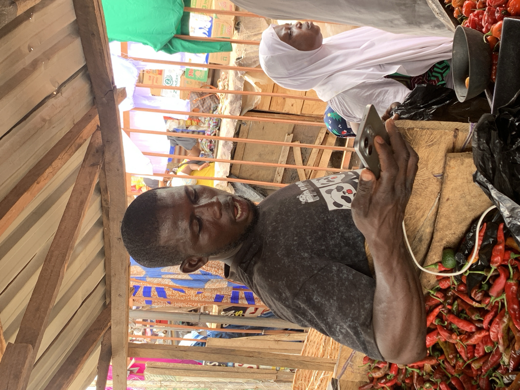 A man is using his smartphone to take a picture or video of a vendor in a market in Ilorin, Nigeria. The vendor, a woman wearing a white hijab, is in the background. The foreground shows the man, a table filled with red peppers and other produce. The market is covered by a corrugated metal roof supported by wooden beams.
