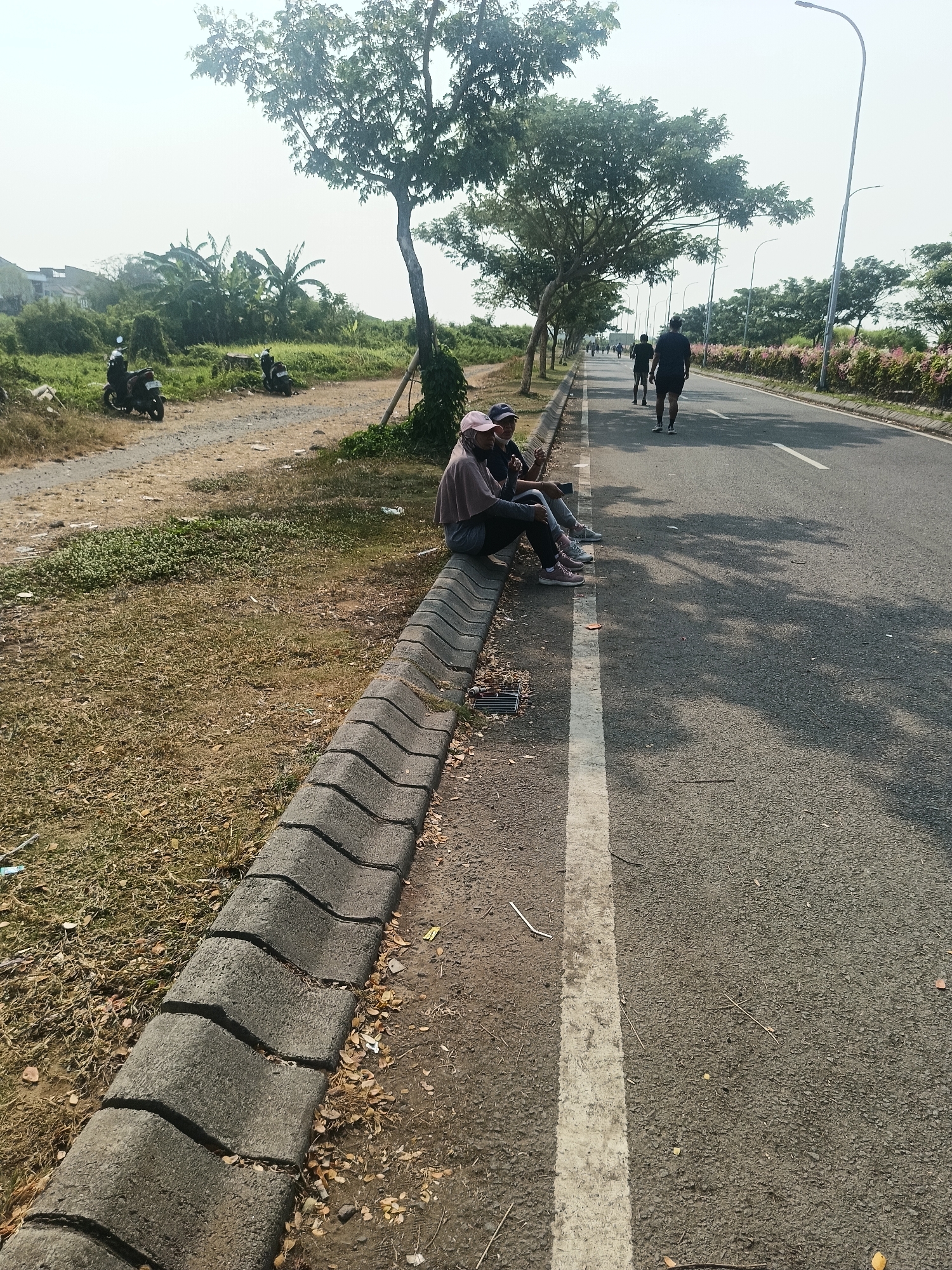 A medium shot captures a sunny daytime scene along a paved road in Punggul, Indonesia. In the foreground, three individuals, two of whom appear to be women wearing headscarves and casual attire, are seated on a concrete curb. They are positioned on the left side of the image, facing the road. To their left is a grassy area with some dry leaves and a gravelly path leading to two parked motorcycles. The road itself, marked with a white line, stretches into the background. Several individuals, seemingly male, are seen running or walking on the road in the middle and far distance, appearing to be engaged in exercise. A line of trees runs along the right side of the road, interspersed with streetlights. The sky is bright and hazy, suggesting a warm day.