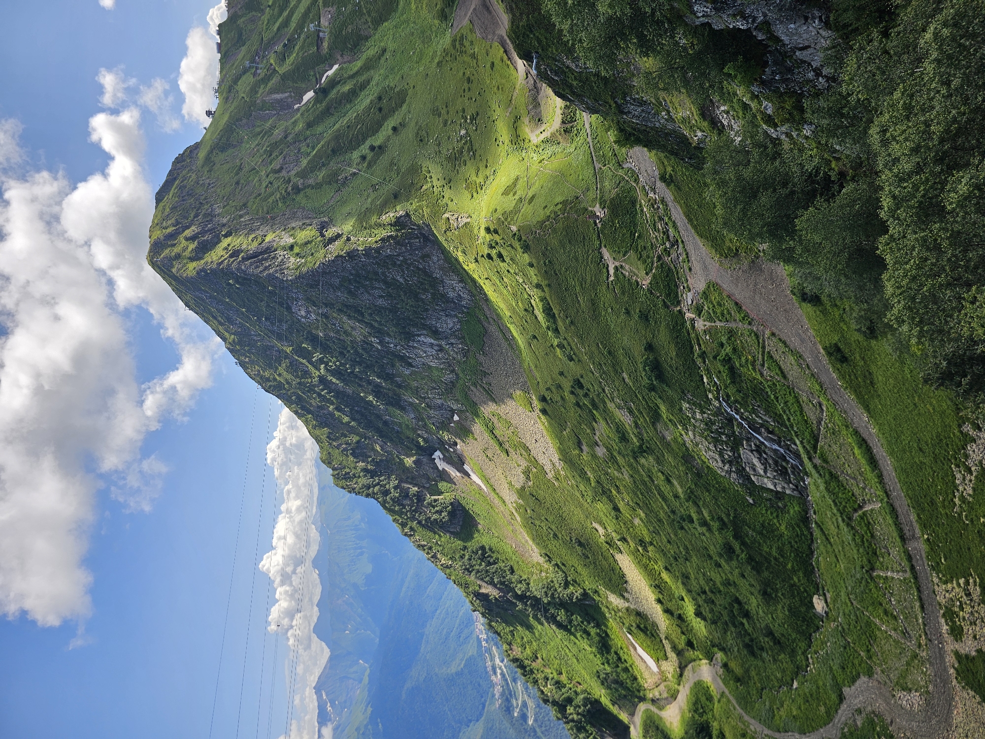 The image depicts the Rosa Khutor ski resort in Estosadok, Russia, on a clear day. A towering, lush green mountain dominates the foreground, its slopes crisscrossed by winding dirt paths and dotted with scattered snow patches. In the distance, a range of hazy blue mountains stretches across the horizon under a sky filled with fluffy white clouds. A few ski lifts and structures are visible on the upper slopes of the mountain, indicating the resort's operational status. The lower part of the mountain reveals a winding road, and a small waterfall cascades down a rocky outcrop. The overall scene is one of natural beauty enhanced by the presence of human infrastructure for recreation.