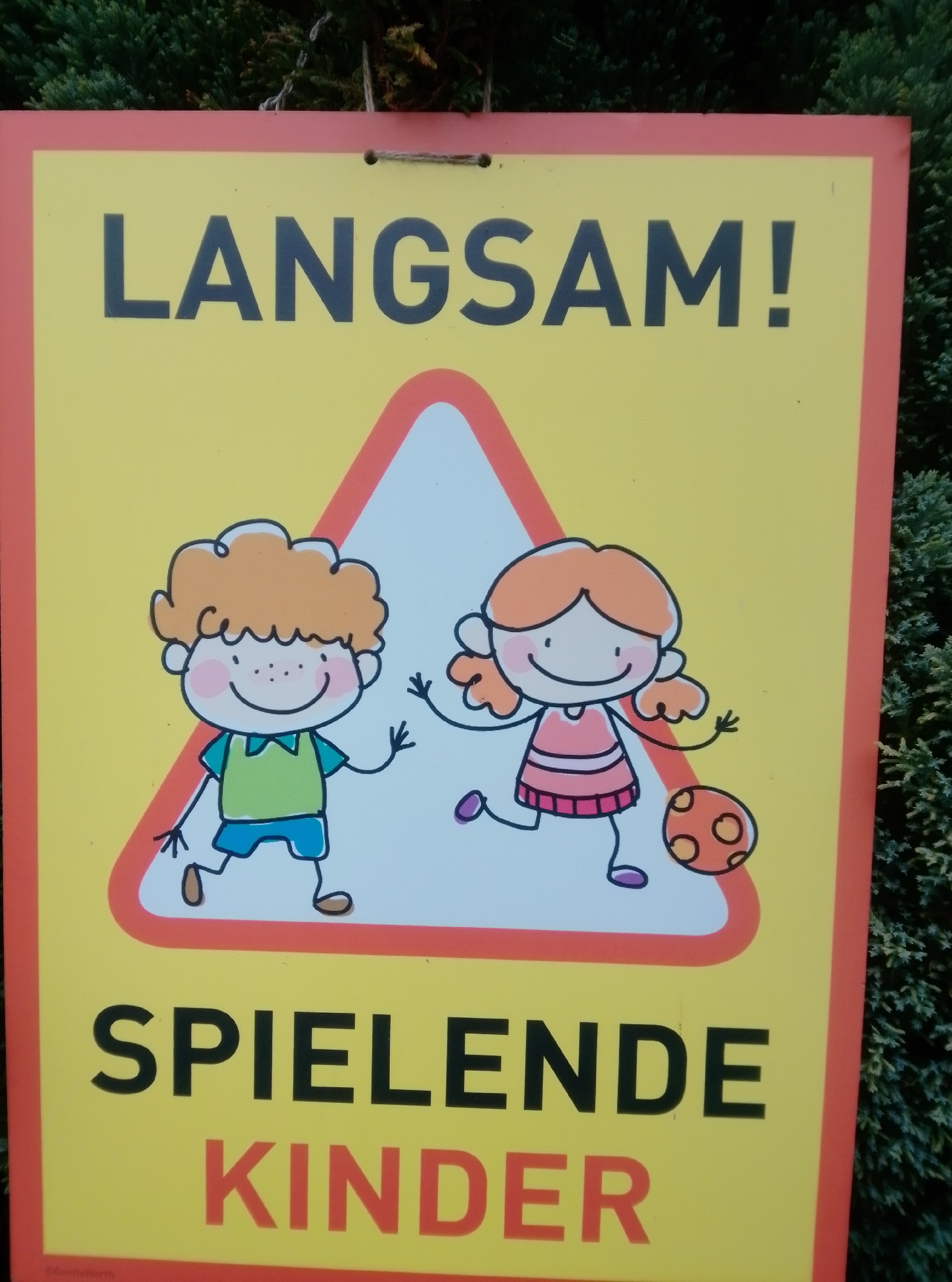 The image shows a yellow sign with red border that reads "LANGSAM!" in large black capital letters at the top. Below this, a red triangle with a white interior contains two cartoon children, one boy and one girl, drawn in a simple, whimsical style. The boy, on the left, has orange curly hair, freckles, a green shirt, and blue shorts, and is waving. The girl, on the right, has orange hair in pigtails, a pink striped dress, and is kicking a soccer ball with orange circles on it. Below the triangle, the word "SPIELENDE" is written in large black capital letters, and underneath that, "KINDER" is written in large red capital letters. The sign is hanging outdoors, in front of a dense green hedge or bush. A small inscription is visible at the bottom left corner, partially obscured. The sign's content, "LANGSAM! SPIELENDE KINDER" (Slow! Playing Children), suggests it is a warning sign to drivers to slow down due to children playing in the vicinity. While the specific location in Wedel, Germany is not visible in the image, such signs are commonly found in residential areas or near playgrounds.