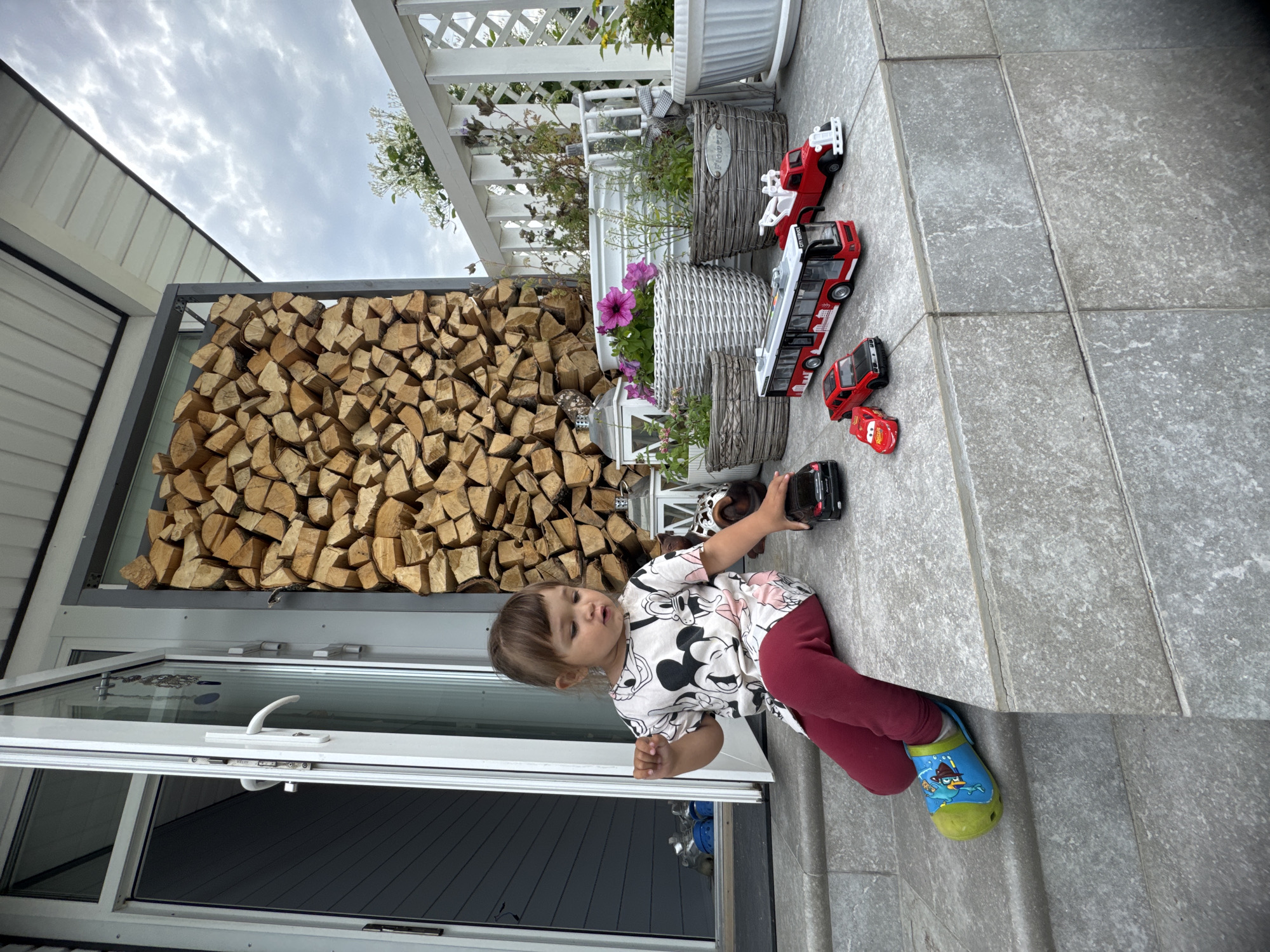 A young girl with brown hair sits on a concrete porch in Oliki, Russia, near an open white door. She is wearing a t-shirt with Mickey Mouse characters and maroon leggings, with a blue and green shoe on her left foot. Scattered around her are several toy vehicles, including a black car, a red car, a red tow truck, and a red and white bus. Behind her, a tall, dark gray metal rack is filled with neatly stacked firewood. Further back, a white railing encloses a balcony with potted plants, some of which have pink and yellow flowers. The scene is set against a cloudy sky.