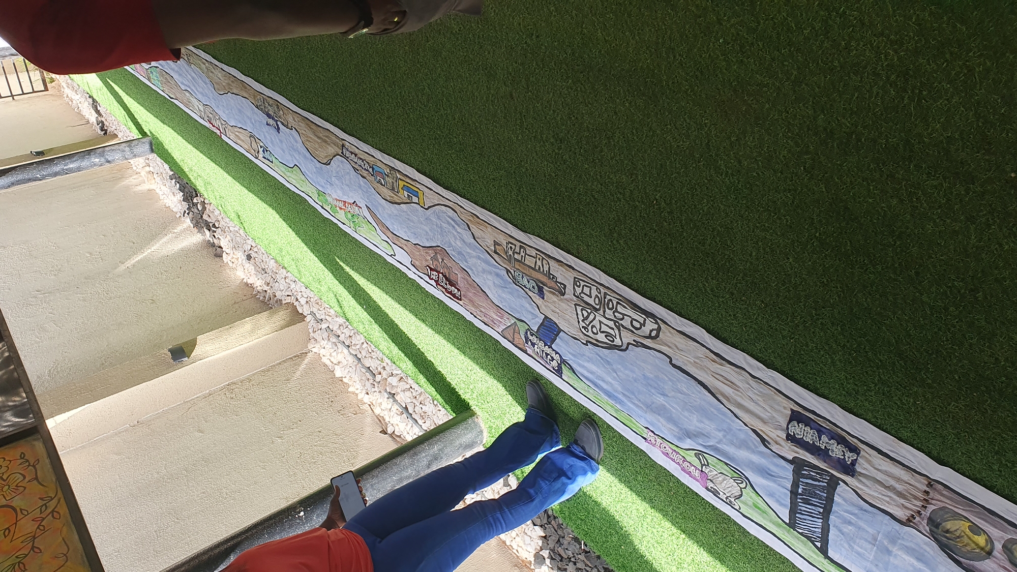 In the city of Unknown, Unknown, a long, unfurled painted banner lies on a green artificial turf surface. The banner depicts a stylized river landscape with various buildings and imagery. To the left, a person wearing a red shirt and blue pants stands, holding a smartphone. The background consists of a light-colored textured wall and a gravel area. The scene is outdoors, with sunlight casting shadows.