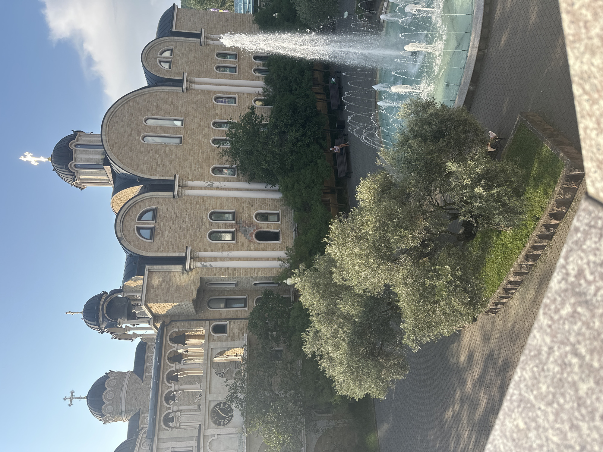 This is an outdoor, daytime, eye-level shot of the Cathedral of the Archangel Michael in Sochi, Russia. The grand stone structure features multiple domes, arches, and windows, with a prominent clock on the left side. In front of the cathedral is a large fountain with jets of water. Several trees with olive-like leaves are in the foreground and middle ground. Two people are visible: one person sitting on a bench near the fountain, and another person reclining on a bench beneath a tree. The ground is paved with interlocking bricks. The sky is blue with some clouds.