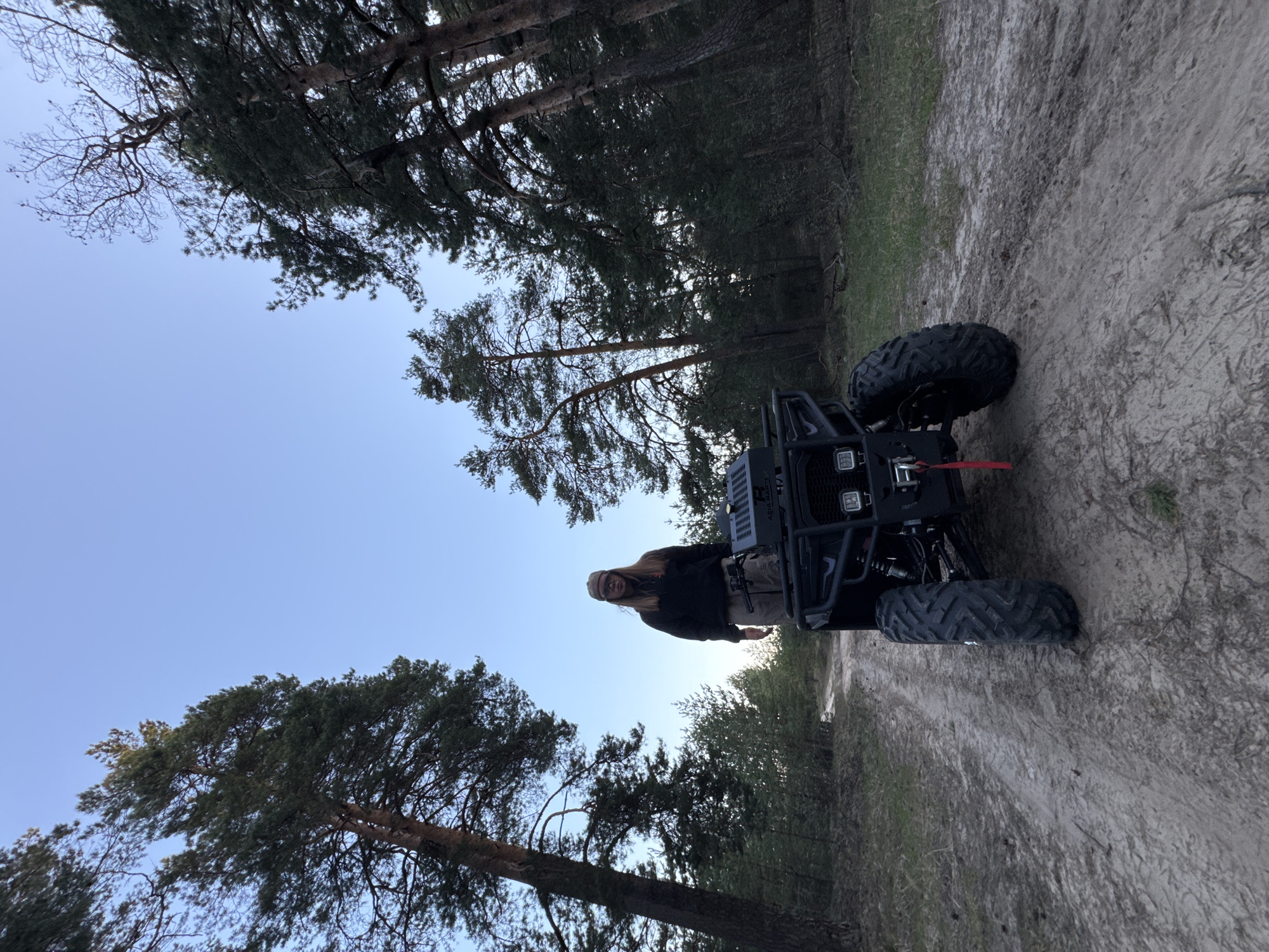 The image shows a person standing next to a black all-terrain vehicle (ATV) on a sandy path surrounded by trees. The location appears to be a wooded area near Bol'shoye Kozino, Russia. The sky is visible above the trees, suggesting it is daytime. The person is wearing dark clothing and a light-colored hat. The ATV is equipped with large tires.