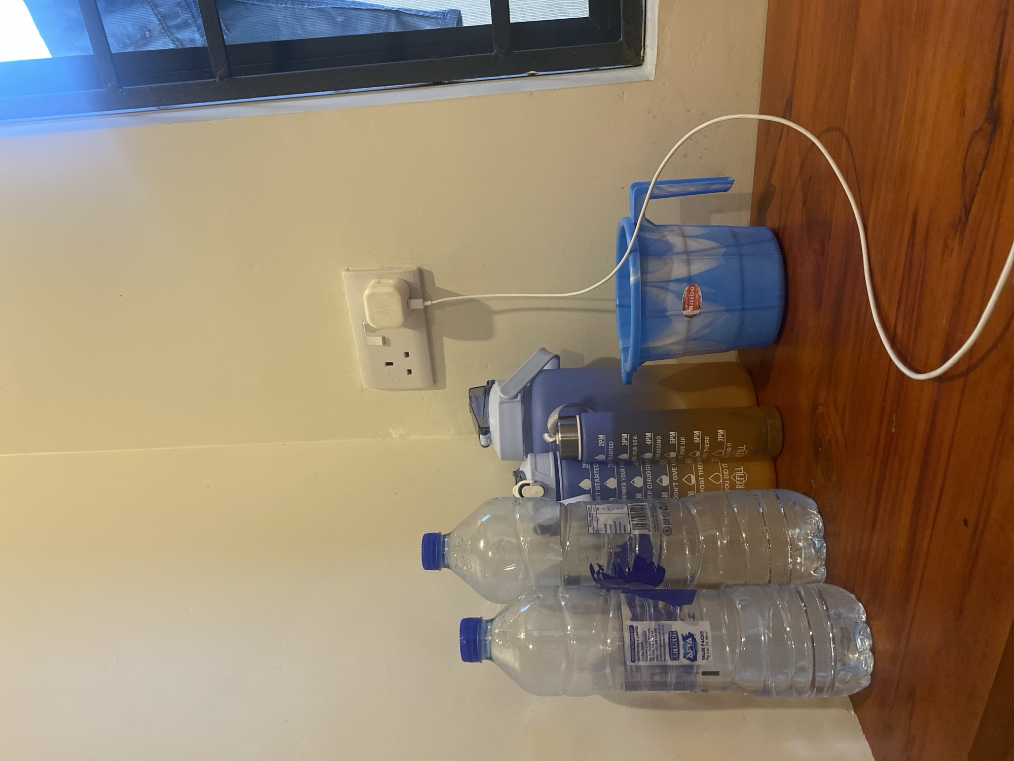 The image displays an indoor scene featuring several water containers and a charging setup, set against a light-colored wall and a wooden floor. A white power outlet with a white adapter is plugged into the wall, from which a white charging cable extends across the floor.
On the wooden floor, a blue plastic mug with a handle, marked "Jumbo," is positioned near the outlet. Below it are two large, graduated water bottles, one blue/purple and the other brown/amber, both featuring time markers (2PM to 7PM) and motivational text including "LET STARTED," "MEMBER YOUR CO," "EP CHUGGIN," "DON'T GIVE UP," "MOST THERE," "YOU DID IT," "REFILL," and "FILL." Two clear plastic water bottles with blue caps are also on the floor, one bearing a label with "AFYA WATERCOM" and "VALUE PACKS," alongside "2P."
A window with a black frame and a mesh screen is visible at the top of the image, suggesting daylight. The power outlet's Type G design is consistent with the provided location context of Dar es-Salaam, Tanzania. The scene presents a collection of items related to hydration and personal electronics in a domestic setting.