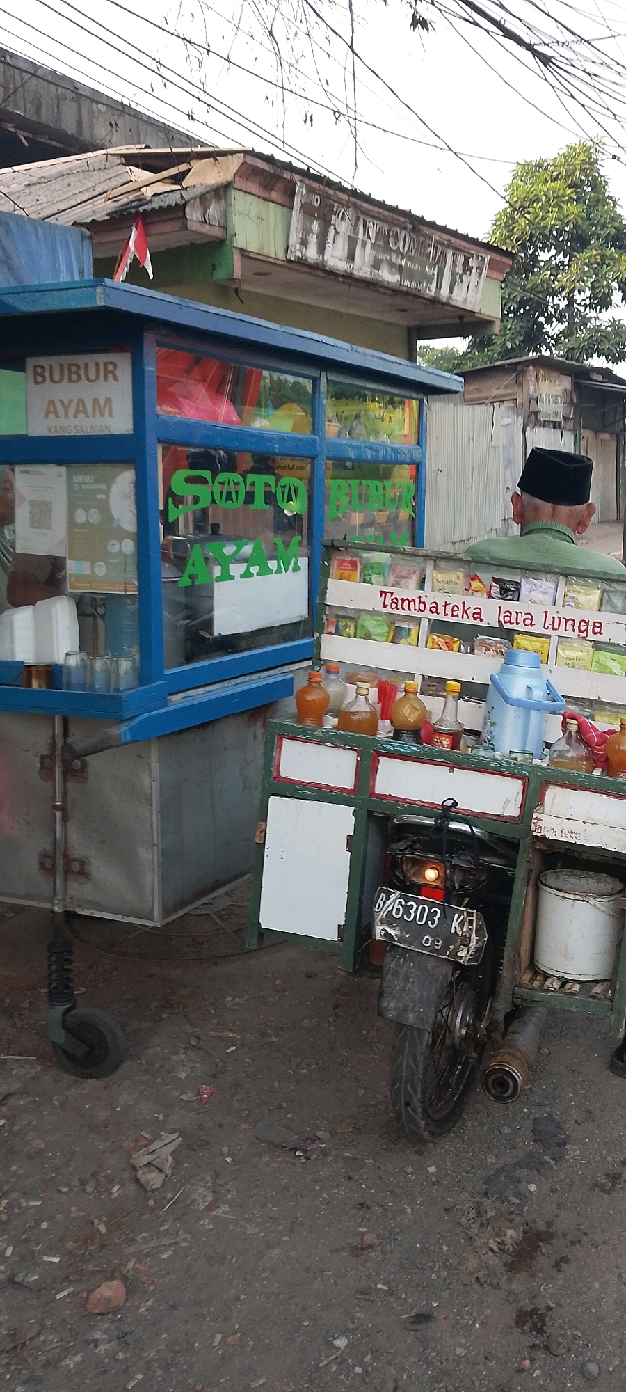 An outdoor street scene in Bekasi, Indonesia, features a food cart selling "Bubur Ayam" (chicken porridge) and "Soto Ayam" (chicken soup). The cart, painted blue, has glass windows displaying menus and food items. A man wearing a green shirt and a black peci (cap) stands behind the cart, facing away from the camera. In front of the cart, there are bottles of condiments and small packages of food items. A motorcycle with a license plate that reads "B 6303 KK" is attached to the cart, suggesting it might be a mobile food business. The surrounding environment appears to be a street with some buildings in the background, showing signs of wear and tear, indicative of a developing urban area. The ground is paved with asphalt and dirt, with some debris scattered around. The overall scene depicts a typical roadside food vendor in an Indonesian city.