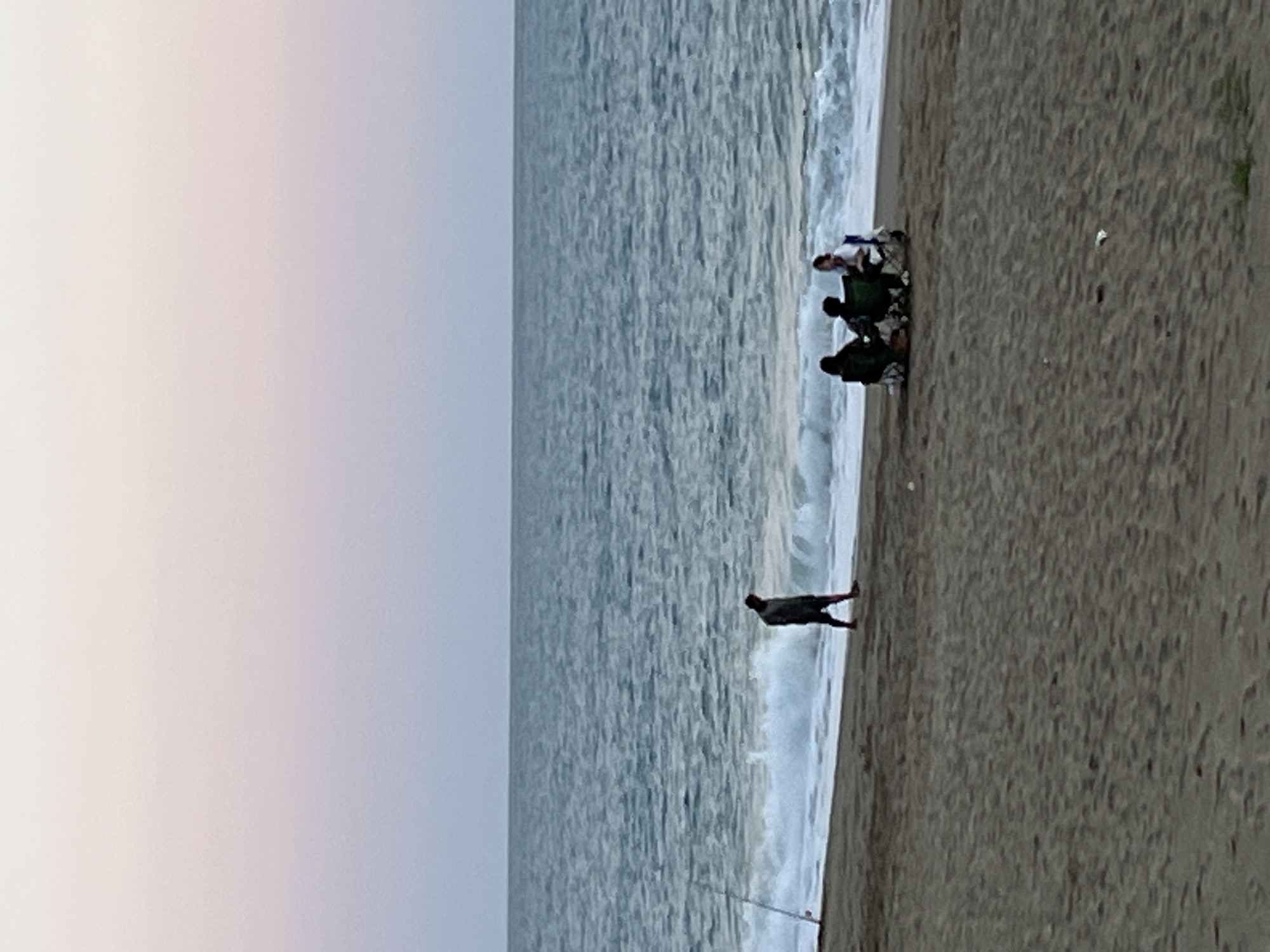 A horizontal view of a beach scene in Alanya, Turkey. The sky is cloudy, transitioning from light pinkish-gray to light gray. The ocean extends from the left side of the frame, taking up the majority of the image. Its surface is textured with light waves.
On the right side of the image is the sandy beach. A group of people are clustered near the shoreline. The people are difficult to identify due to the distance and resolution. One individual is walking near the water's edge. A fishing pole is partially visible in the sand near the bottom of the frame.