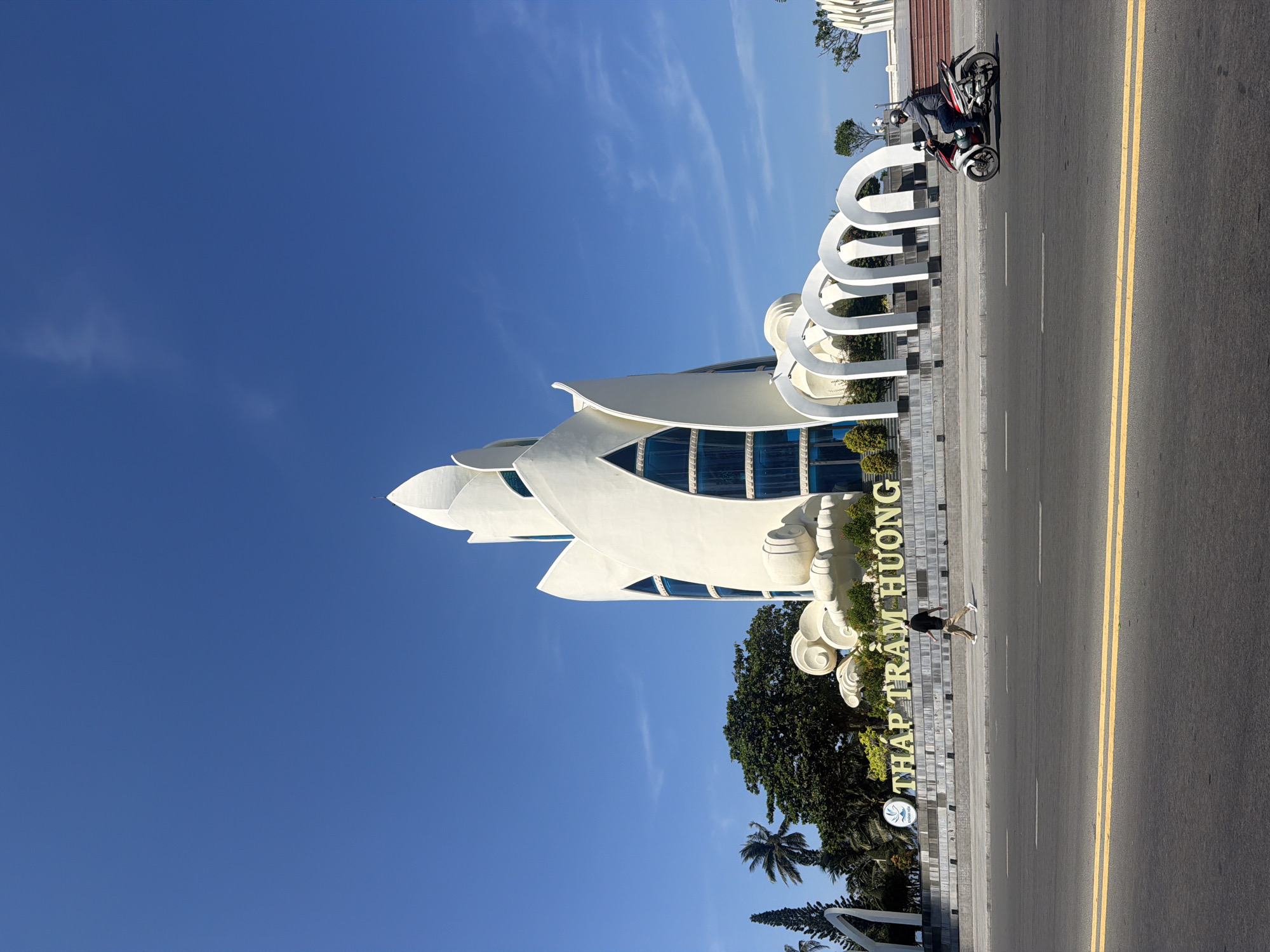 The image depicts a frontal view of the Po Nagar Cham Towers, a prominent landmark in Nha Trang, Vietnam. The towers, built in a distinctive white lotus-flower design with blue-tinted windows, rise majestically against a clear blue sky. At the base of the towers, a stone monument reads "THÁP TRẦM HƯƠNG" in large, yellow, three-dimensional letters, with a smaller emblem to the left.
In the foreground, a paved road with double yellow lines runs horizontally across the image. A man wearing a dark t-shirt and beige pants is walking on the sidewalk to the left of the road. To the right, a person is riding a scooter, also heading towards the right side of the frame.
Behind the monument, lush green trees and palm trees provide a verdant backdrop. The architecture of the towers features intricate curves and layered petal-like structures, culminating in pointed spires at the top. To the right of the main structure, a series of white arched colonnades are visible. The overall scene conveys a bright, sunny day in a coastal city.