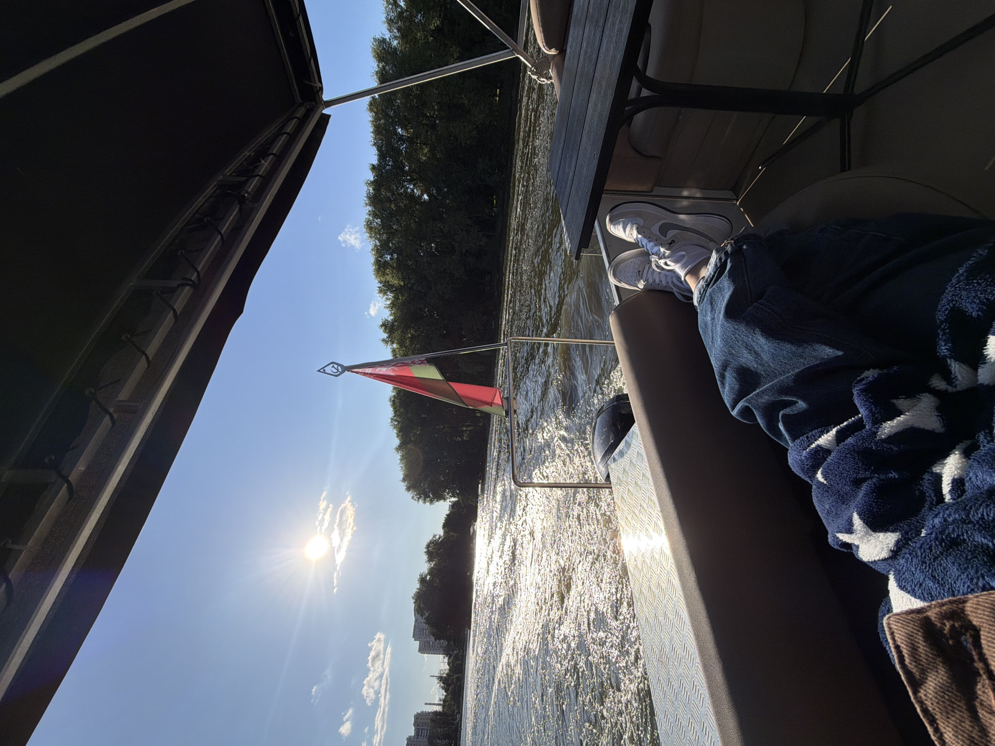 The image depicts a person relaxing on a boat sailing on the Svislach River in Minsk, Belarus. The person's feet, clad in white sneakers and blue jeans, are resting on the seat, covered partly by a blue blanket with white stars. They are positioned near a dark-colored table. The boat moves through a body of water, creating ripples and reflections of the bright sun. Trees line the riverbank in the background, with buildings visible in the distance on the left. A flag, red and green, flutters on a pole at the rear of the boat. The sky is clear blue with a few scattered clouds.