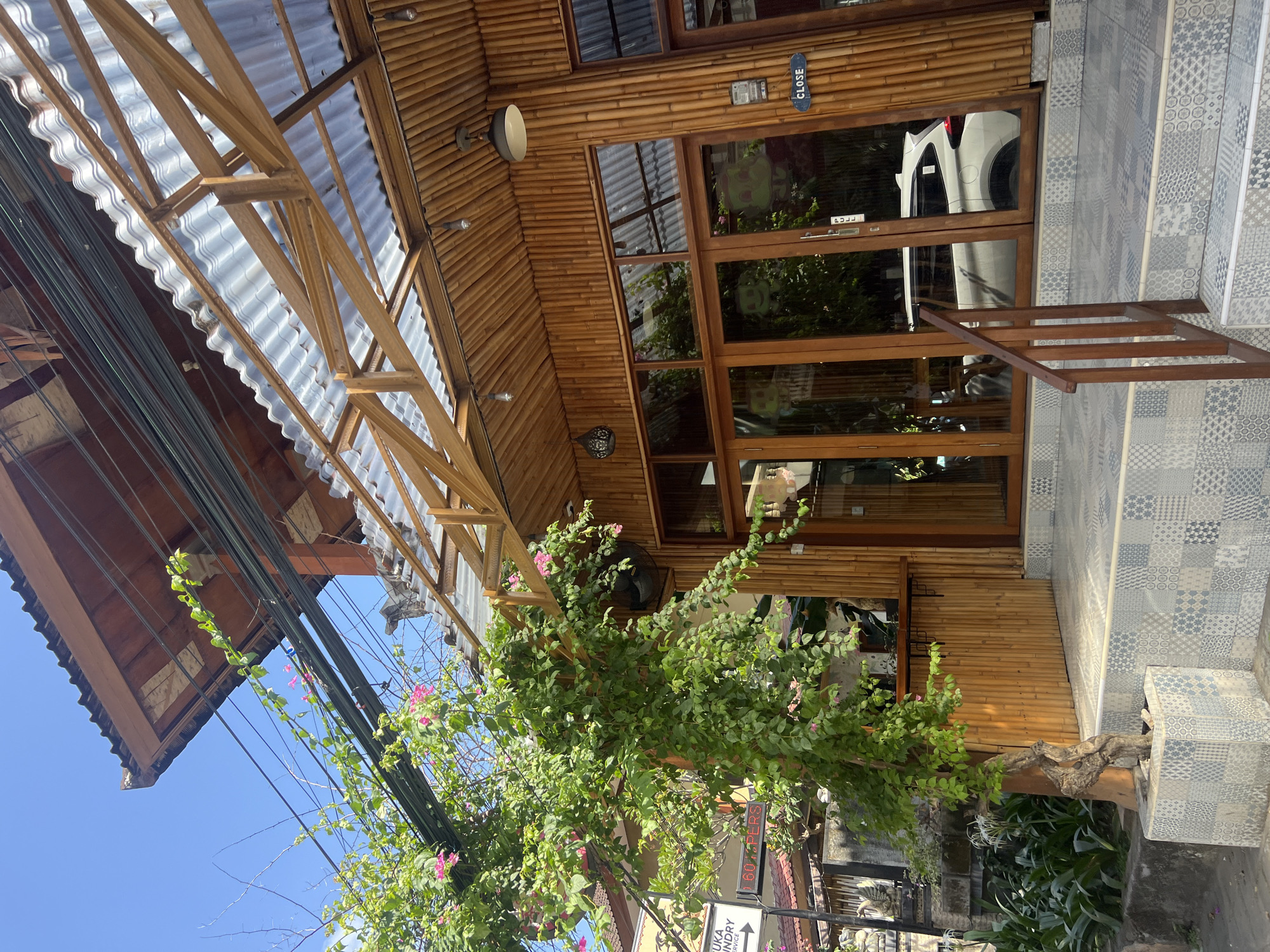 The image depicts the exterior of a building with a bamboo facade and a wooden and corrugated metal roof in Kuta, Indonesia. A large tree with green leaves and pink flowers is in the foreground, growing from a patterned planter. The building has large glass doors and windows, reflecting the surrounding environment. A white car is visible through the glass doors. A sign reading "CLOSE" is attached to the bamboo wall. The pavement in front of the building is tiled with a mosaic of blue and white geometric patterns. Overhead, electrical wires are strung across the scene. The overall impression is a tropical setting with a rustic and natural architectural style.