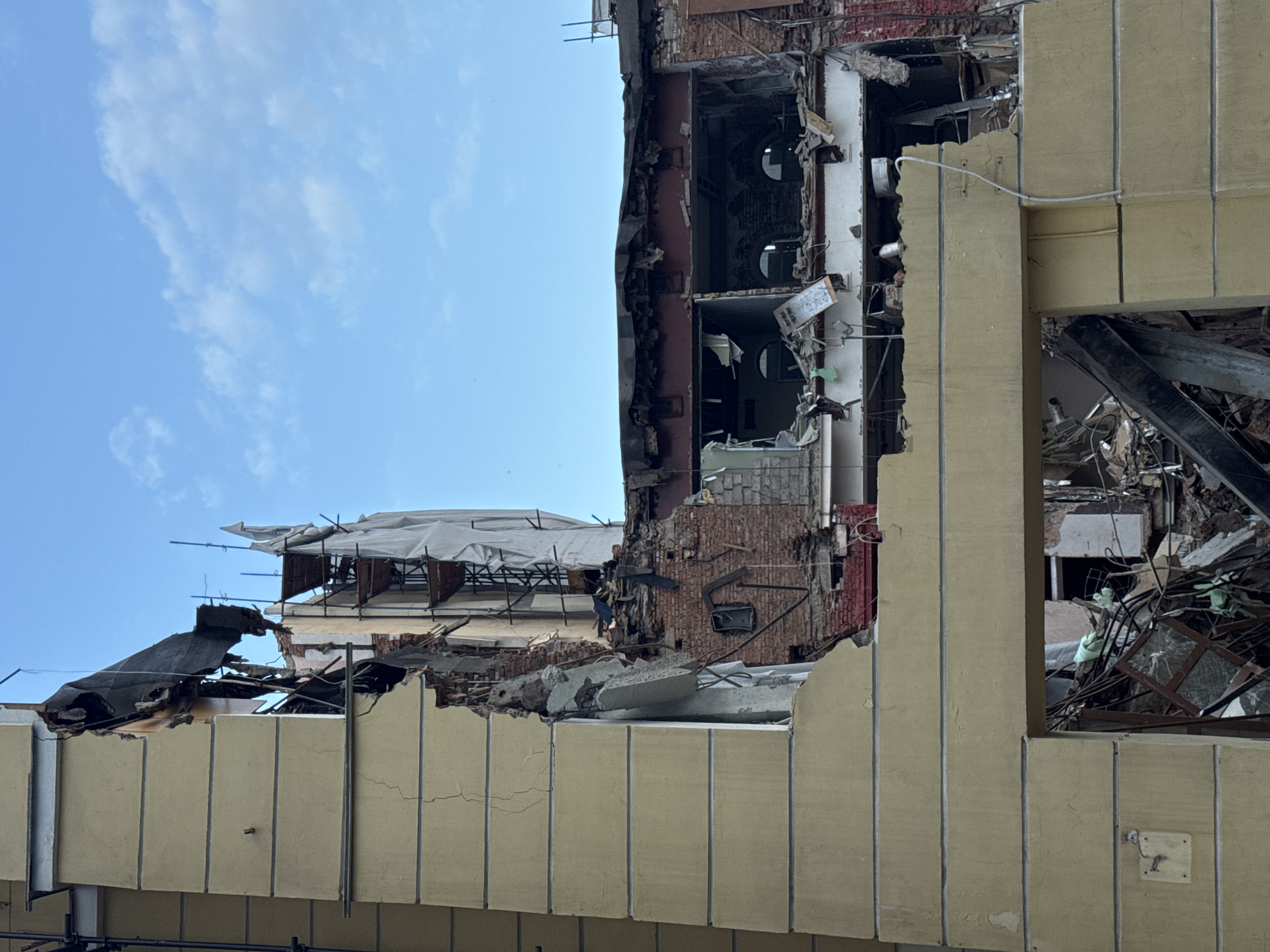 A severely damaged building stands in Moscow, Russia, under a partially cloudy sky. The building's façade is crumbling, exposing brickwork, torn walls, and structural debris. One section is covered with a protective tarp and scaffolding. The destruction appears extensive, suggesting a possible collapse or explosion. The scene lacks signs of human activity.