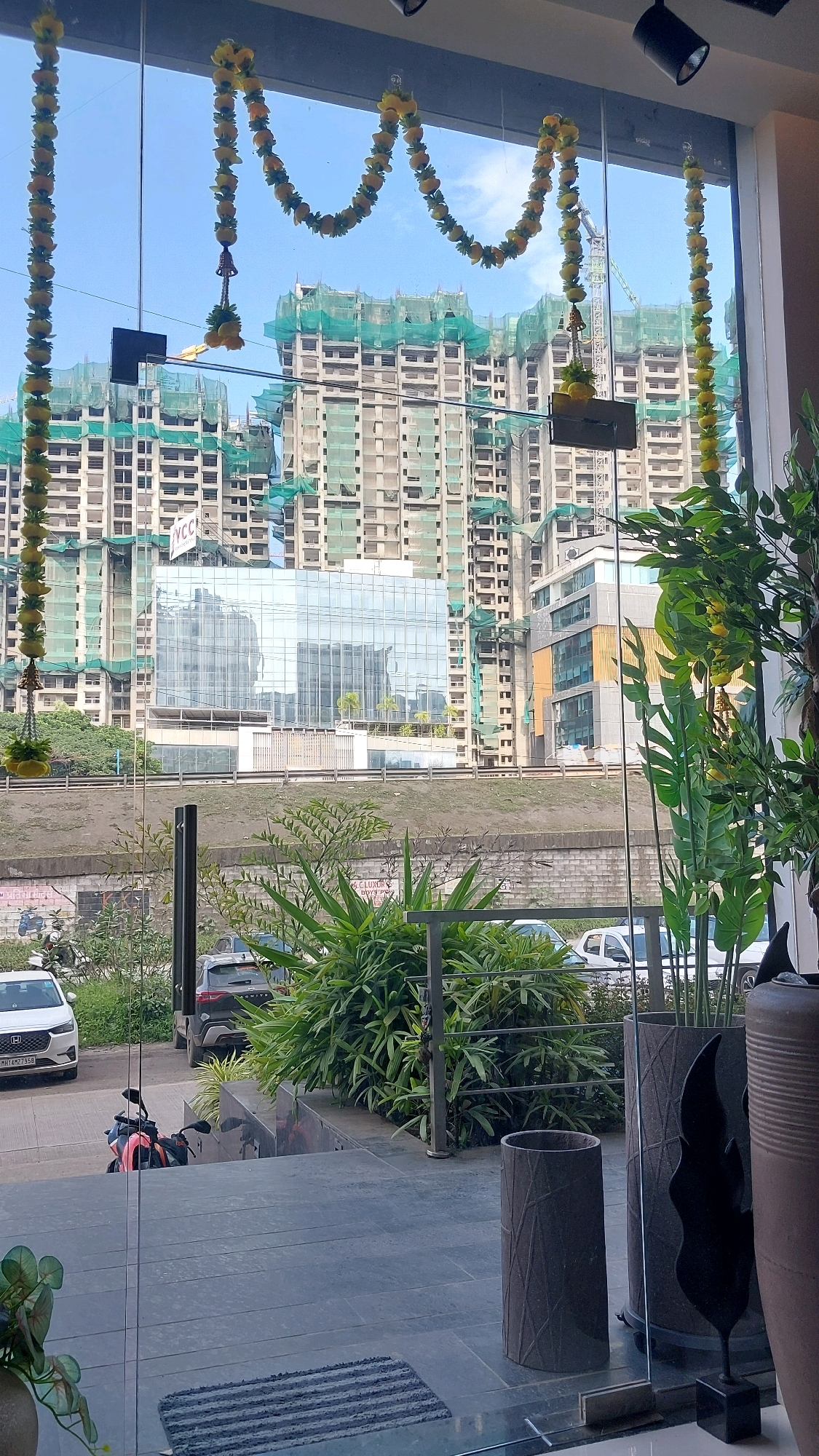 The image depicts a view through a large glass window or door, looking out at a cityscape under construction. In the foreground, part of a modern interior is visible, with large dark-tiled flooring and decorative planters. A striped doormat is positioned on the floor. Outside, a row of green plants and trees forms a barrier between the building and a road where several cars and a motorcycle are parked.
Beyond the immediate surroundings, numerous high-rise buildings are under construction, clad in scaffolding and green netting. One building in the mid-ground features a reflective glass facade. In the distance, a construction crane is visible against a clear blue sky.
The yellow flower garlands strung across the top of the window suggest a festive or celebratory occasion, possibly related to the building's completion or a holiday. The overall scene conveys a sense of urban development and ongoing progress in Pune, India.