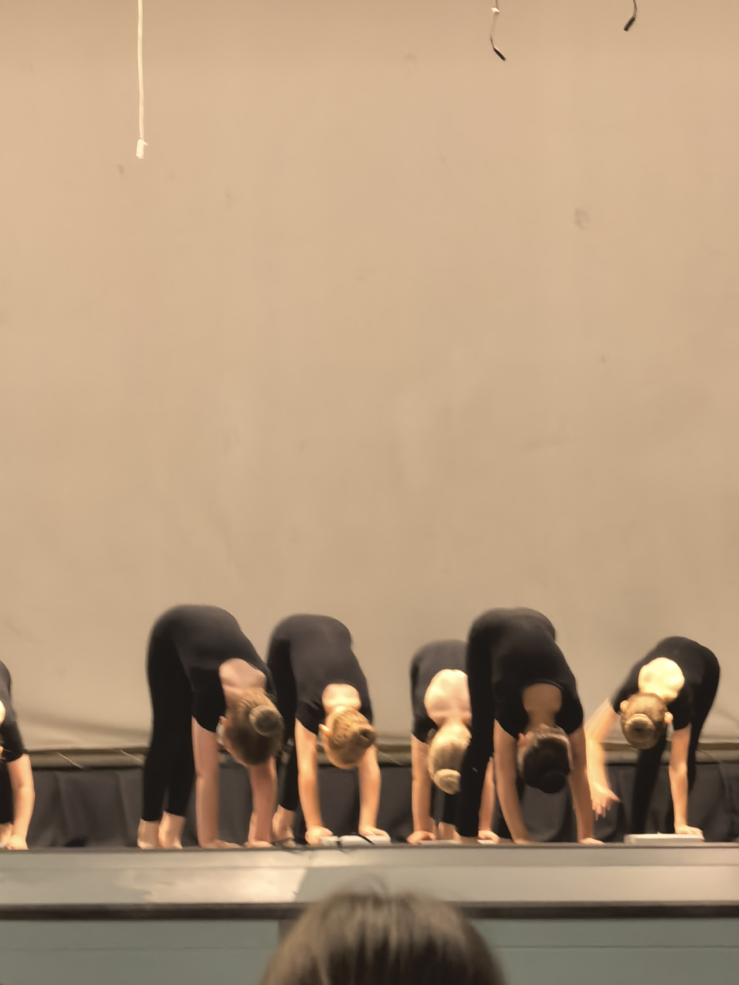 The image shows a group of young girls in black leotards and tights performing a dance routine on a stage. They are all bending forward with their heads down, hands on the floor in front of them, and legs straight. The girls have their hair in buns. The stage has a dark curtain behind the dancers and a light-colored backdrop. The lighting on the stage is a warm yellow hue. The overall impression is of a dance rehearsal or performance.