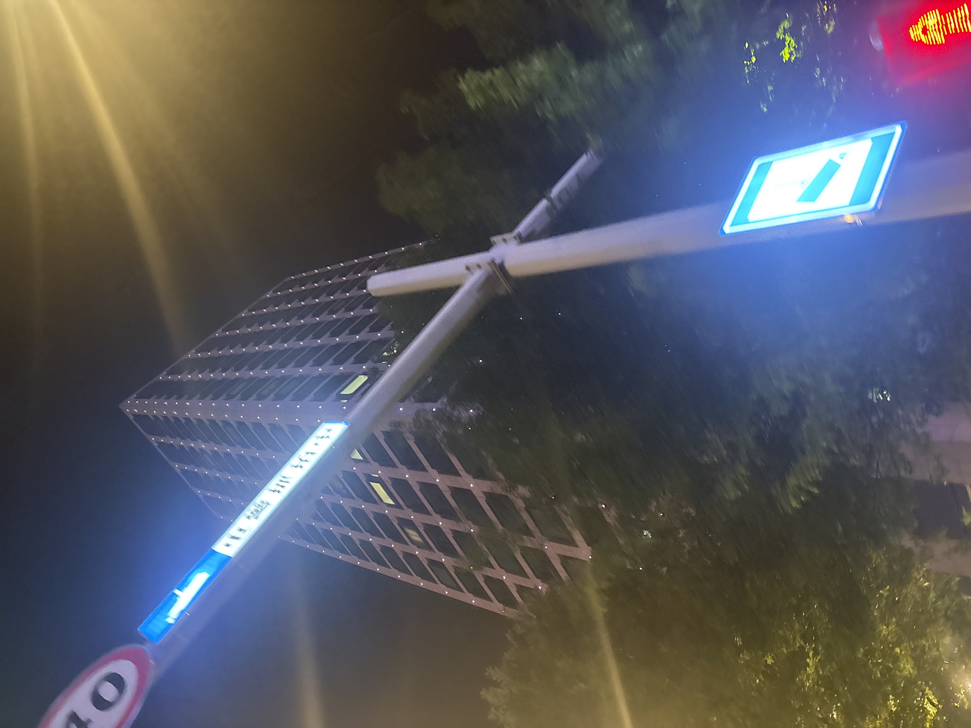 The image shows a street scene at night in Suzhou Industrial Park, China. A tall, modern building with illuminated windows dominates the background. In the foreground, a street light pole is visible, adorned with several signs. One sign, at the top left, appears to be a speed limit sign, with the number "40" visible. Below this, another sign is illuminated in blue and displays some text. Further down the pole, a blue and white diamond-shaped sign with an arrow pointing to the left is also illuminated. The surrounding area is dark, with trees partially obscuring the building, and streetlights casting a yellow glow. There are no people or vehicles clearly visible in the frame.