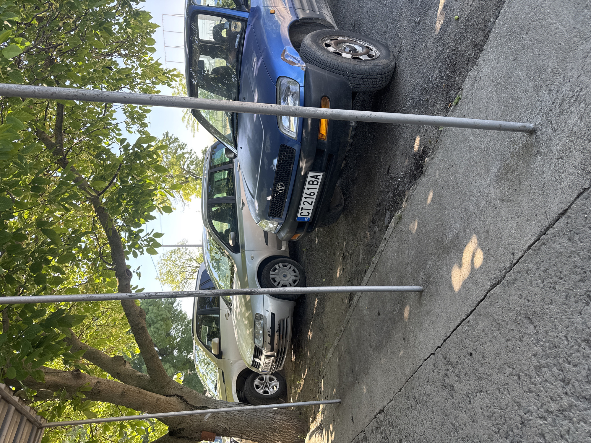 The image shows two parked cars on a street in Bulgaria, potentially in or near Sarnevo. The first car is a blue Toyota RAV4 with a license plate reading "CT 2161 BA." The second car is a silver Fiat Stilo, its license plate reads "CT8647AI". The cars are parked adjacent to a concrete sidewalk next to a tree and a metal railing or fence, casting shadows on the concrete. The vehicles appear to be in a residential or urban environment, with tree cover suggesting possible parkland nearby.