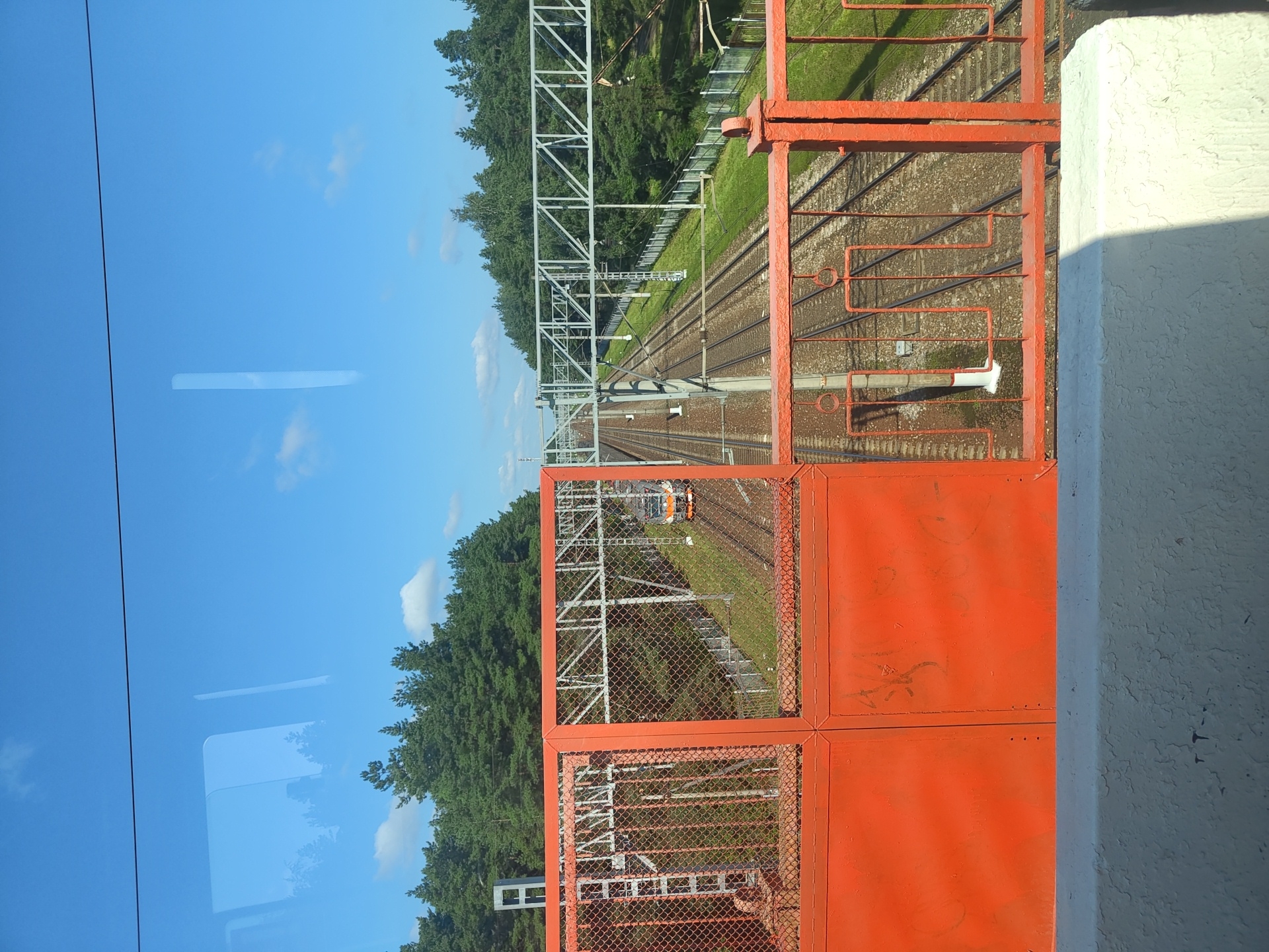 The image captures a view of a railway line from an elevated perspective, likely a pedestrian overpass. In the foreground, a bright orange metal fence and gate dominate the lower half of the frame, with visible graffiti on the gate's panels. The fence is made of mesh and has decorative circular and rectangular elements. Beyond the fence, multiple railway tracks stretch into the distance, lined by green grass and trees. A train with an orange and gray color scheme is visible on one of the tracks, moving away from the viewer. Overhead, electrical catenary lines are suspended above the tracks. The sky is a clear blue with a few white clouds. The overall scene depicts a functional railway infrastructure surrounded by natural elements, under a bright sky. This location is identified as being in Ramenskoye, Russia.