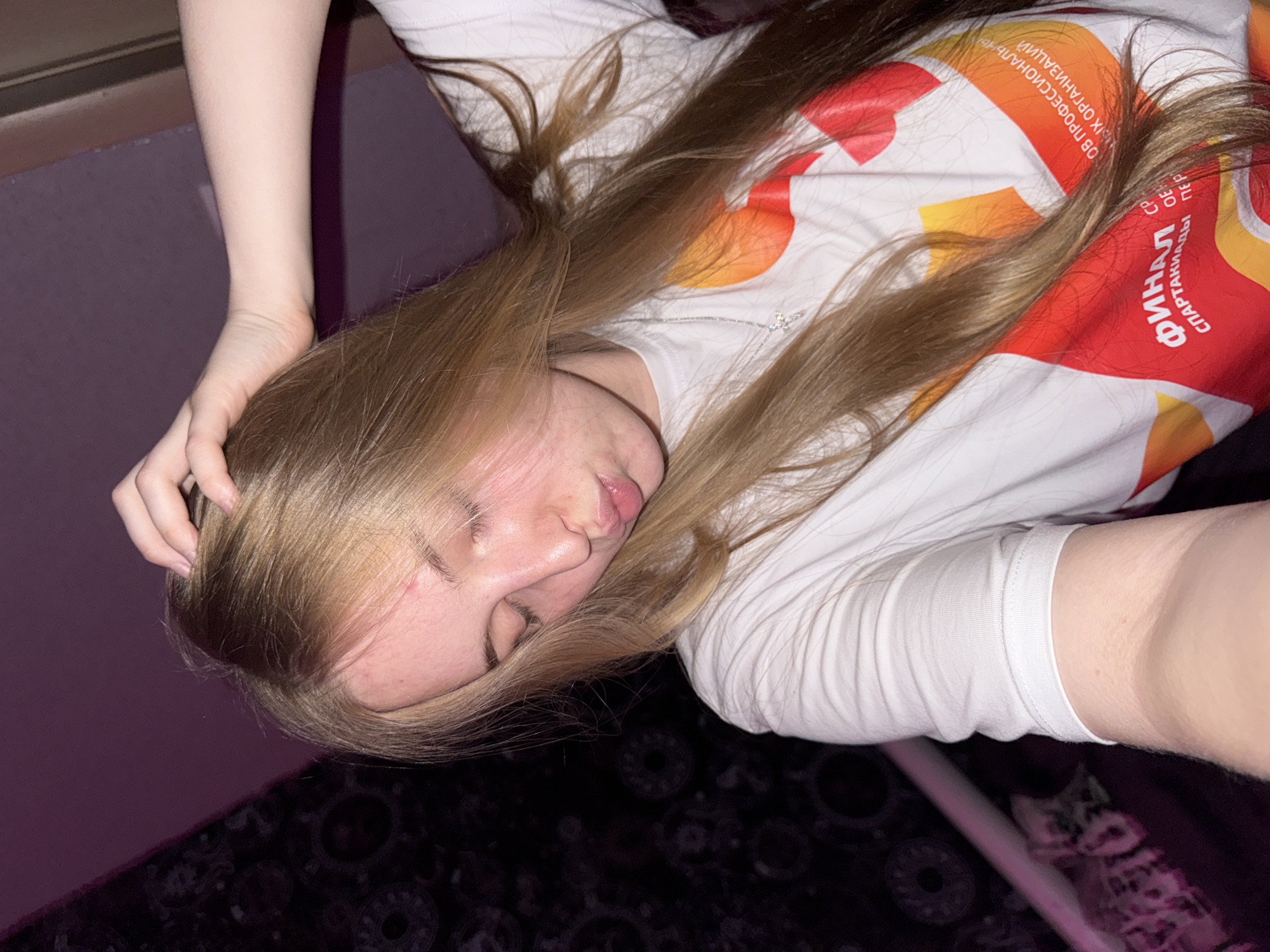 A young woman with long blonde hair is captured in a close-up, self-portrait, likely taken indoors. She is wearing a white t-shirt with a colorful graphic featuring red, orange, and yellow elements and text in Russian. Her eyes are closed, and her lips are puckered in a kiss pose. Her right hand is raised, with fingers splayed through her hair near the crown of her head. The lighting in the scene is dim and casts a purplish hue, suggesting it may be nighttime or the use of colored lighting. A dark patterned curtain or fabric is visible in the background to the left, and a white cylindrical object, possibly a pole or pipe, is partially visible on the lower left. The overall ambiance is casual and personal. There is no indication of location in a city.