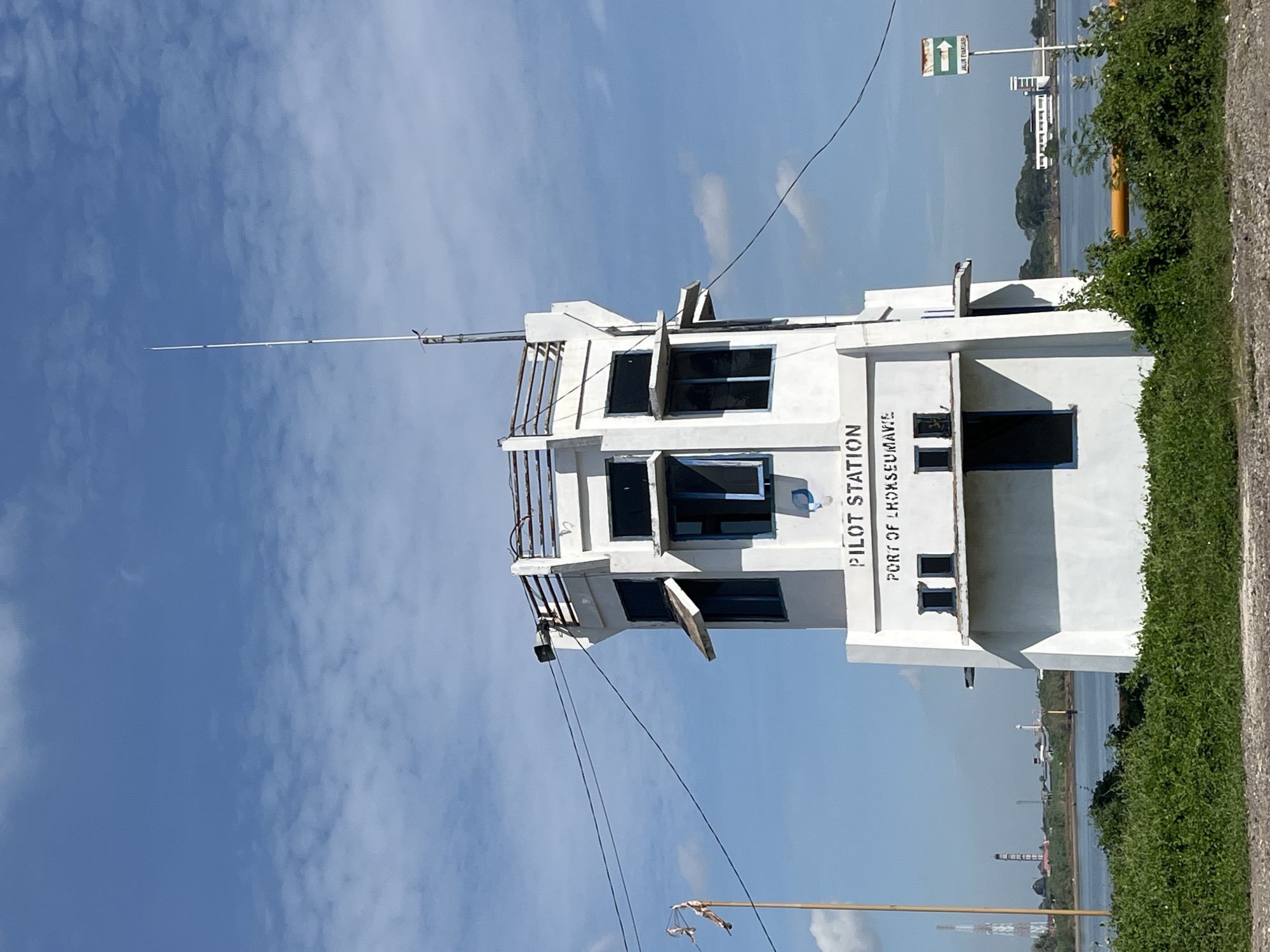 The image shows a white pilot station building, identified as "PILOT STATION, PORT OF LHOKSEUMAWE," against a partly cloudy blue sky. The structure has multiple levels with dark-framed windows, some of which are partially open. A tall, thin antenna extends from the top of the building. Communication wires are visible. The building is located near a body of water with some foliage and infrastructure visible in the distance. The photo was taken in Tambonbaroh, Indonesia. Green vegetation is visible on the right side of the image, bordering a road or path. A directional sign is located near the building.
