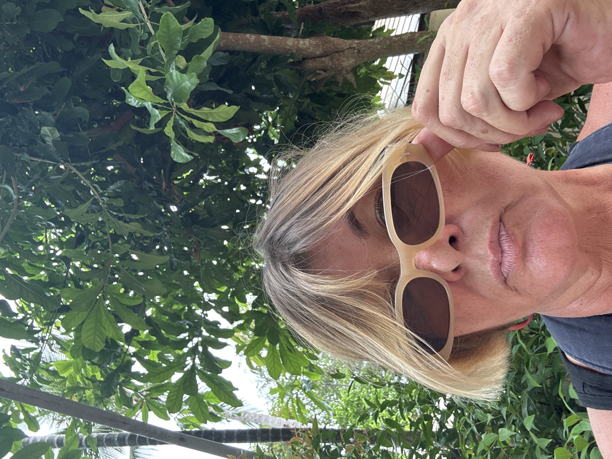 A close-up, low-angle selfie shows a fair-skinned person with blonde hair and sunglasses. They are holding the sunglasses up to their face with their right hand. The person is wearing a dark-colored shirt. The background is lush and green, with large leaves of trees and branches visible. There are hints of a white structure, possibly a fence or building, behind the foliage. The lighting suggests it is daytime, and the vibrant greenery implies an outdoor, possibly tropical, location. The person's expression is partially obscured by the sunglasses and hand, but their lips are slightly parted. The overall mood is casual and relaxed. There is no visible text in the image.
